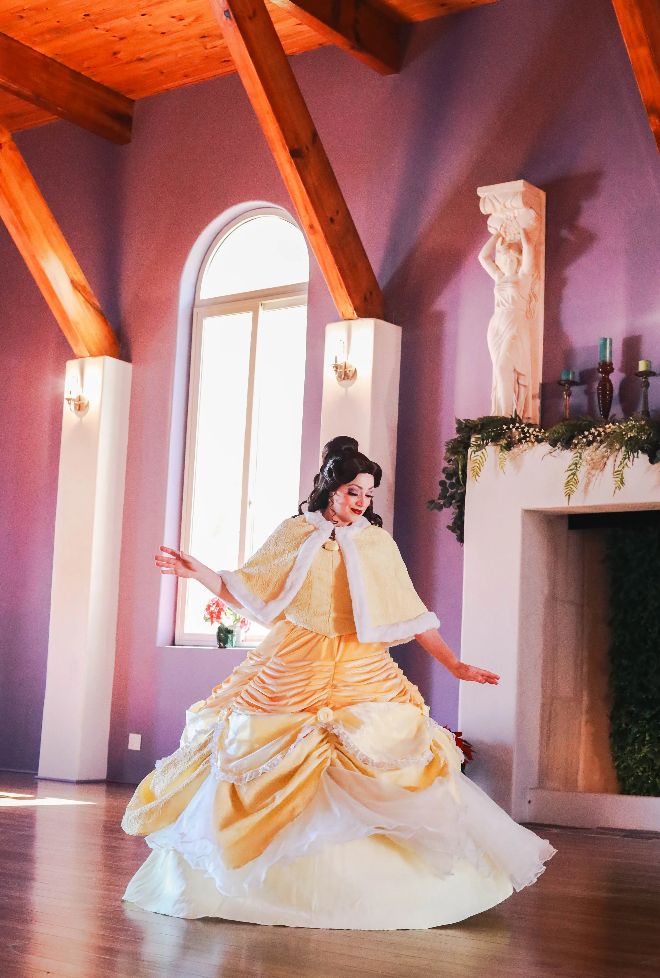 A woman dressed as is dancing in a purple-walled room with wooden beams and a large window. She wears a yellow ball gown with a swept-up hairstyle and a matching yellow cape. There is a white sculpture of a woman on a shelf near the fireplace, decor