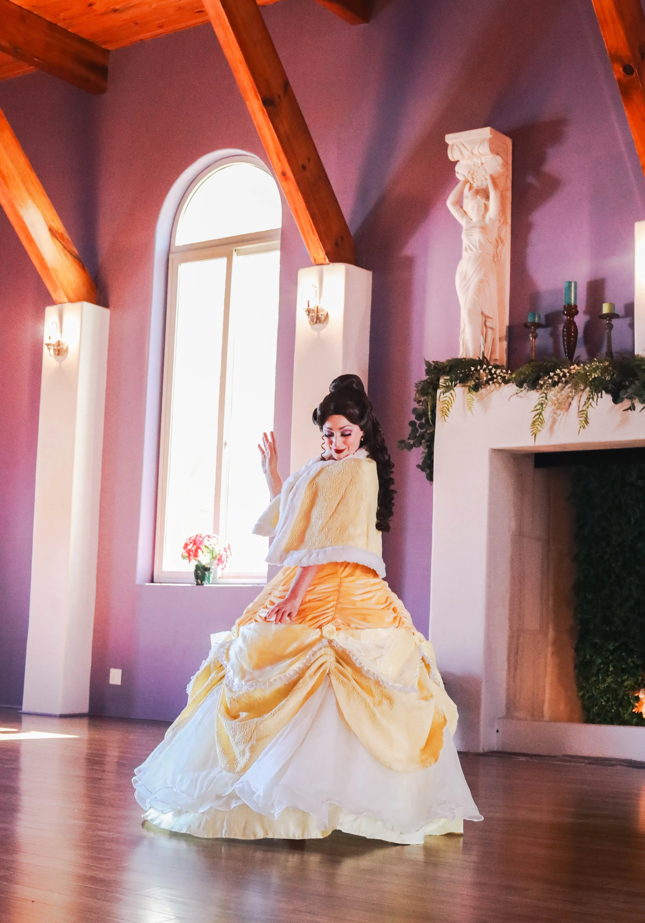A woman dressed in a yellow and white vintage-style gown, posing in a decorated room with purple walls, wooden beams, and a large window with sunlight, an ornate white statue on the fireplace mantel, and greenery.