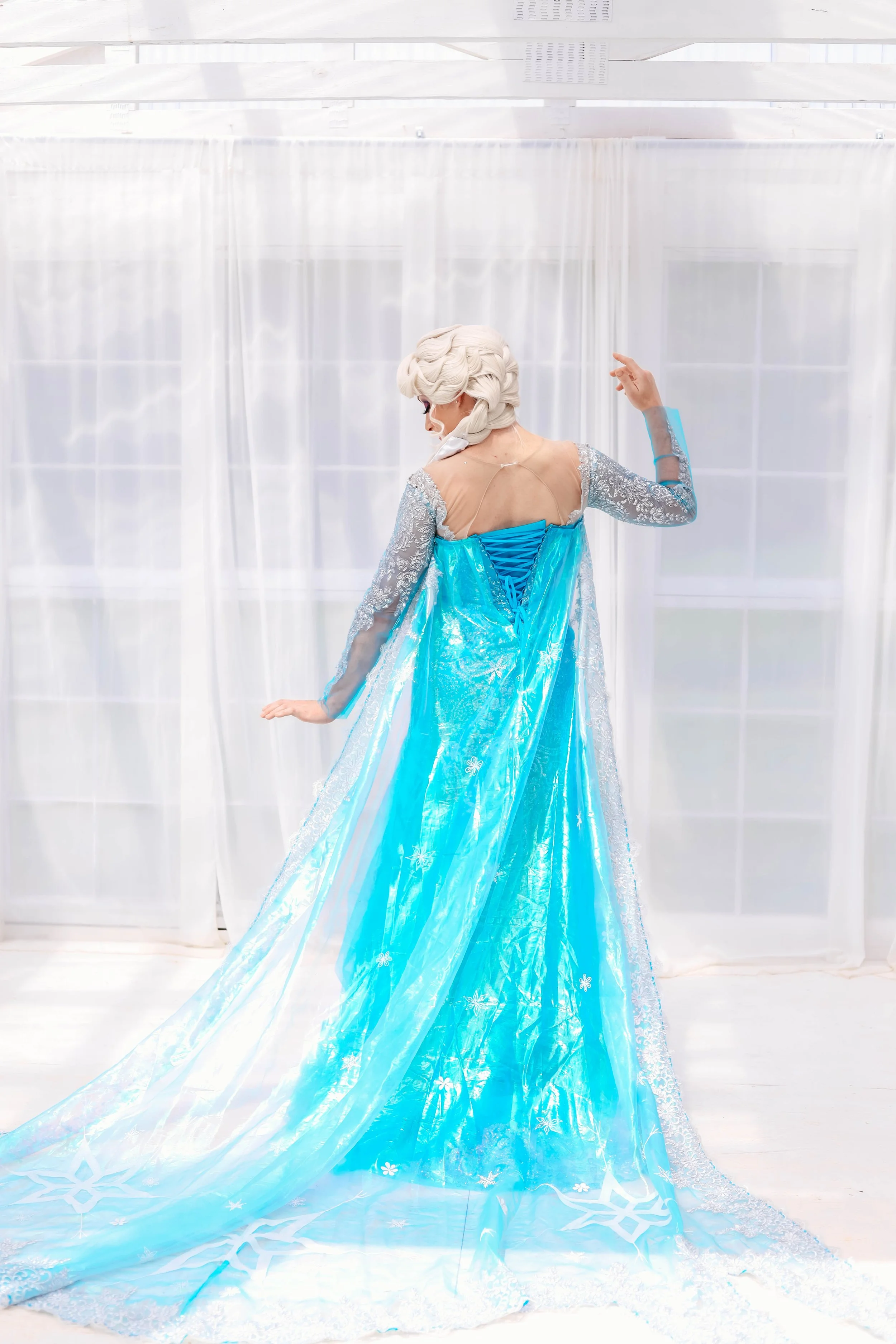 A person dressed as the ice queen, wearing a light blue, sparkly dress with a flowing cape, standing in front of white curtains.