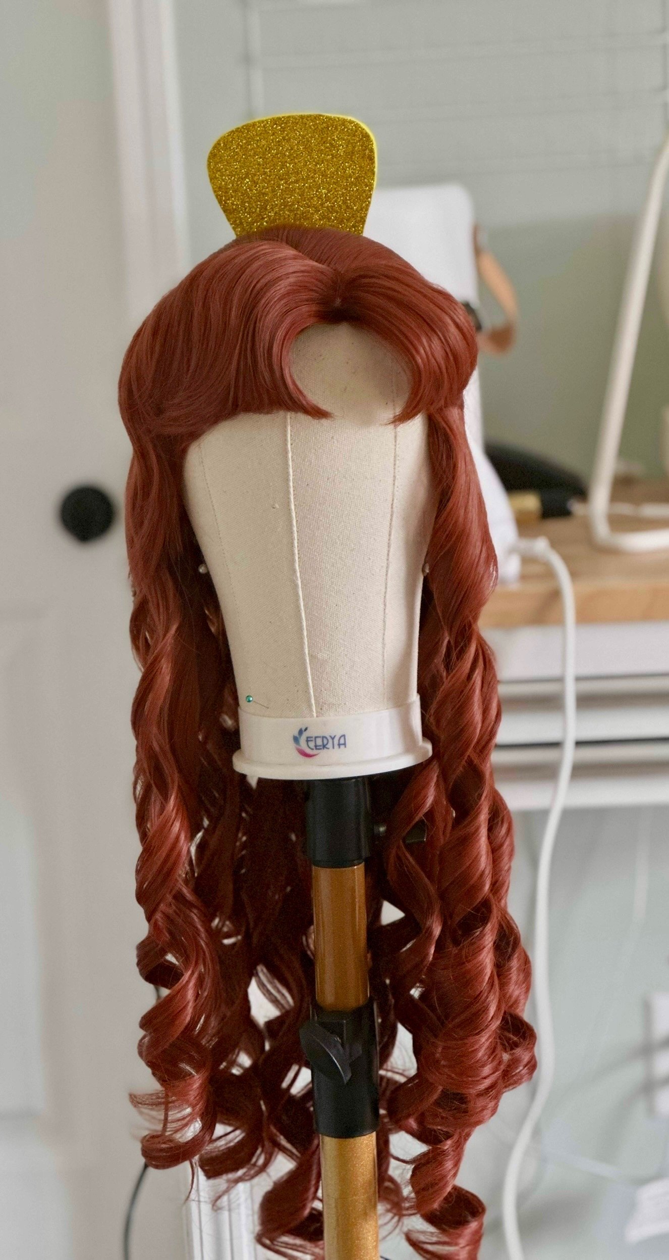 A red wig with long, curly hair and a glittery yellow hair accessory, mounted on a wig stand, in a room with a wooden table and white wall in the background.