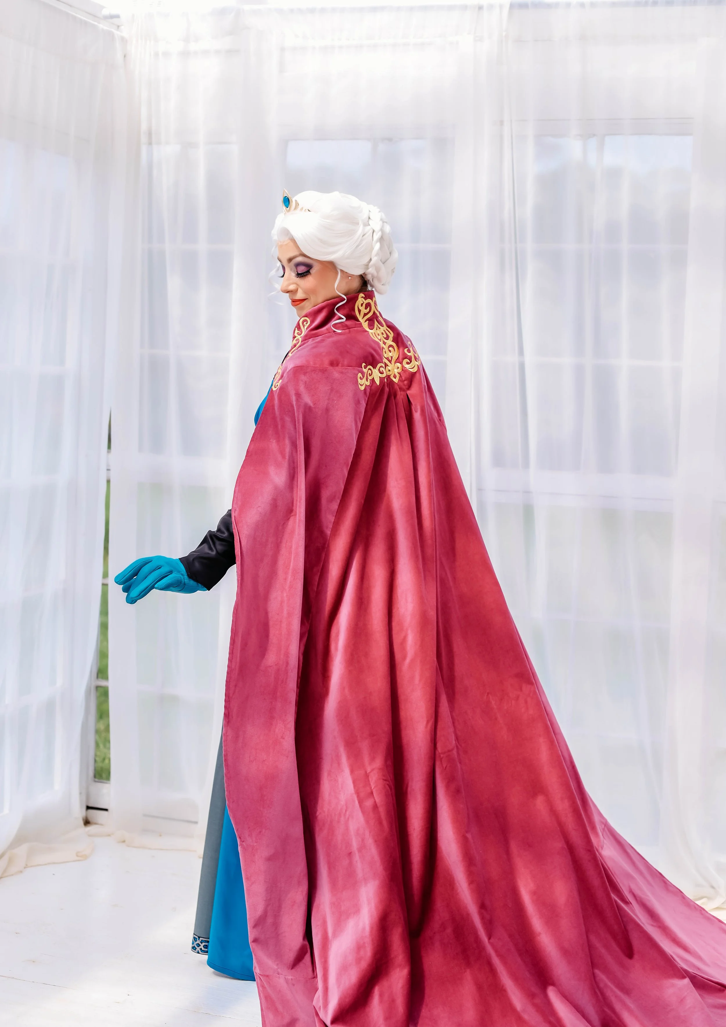A woman dressed as the ice queen, wearing a long blue gown and a red cape with gold embroidery, standing inside a white, curtain-enclosed space.
