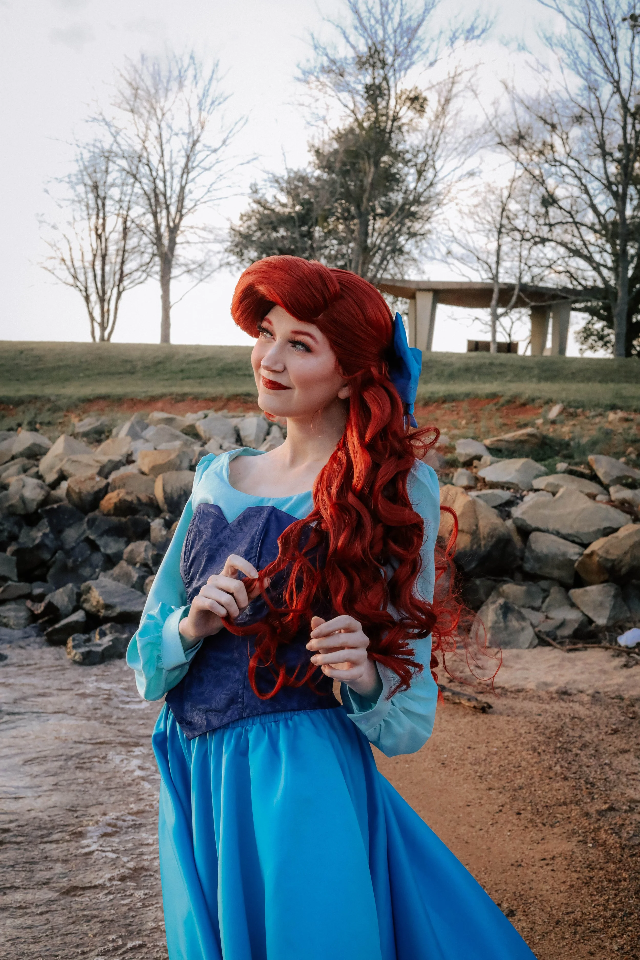 A woman dressed as a princess with long red curly hair, wearing a blue dress with matching bow in her hair, standing outdoors on a rocky beach with trees and a pavilion in the background during daylight.
