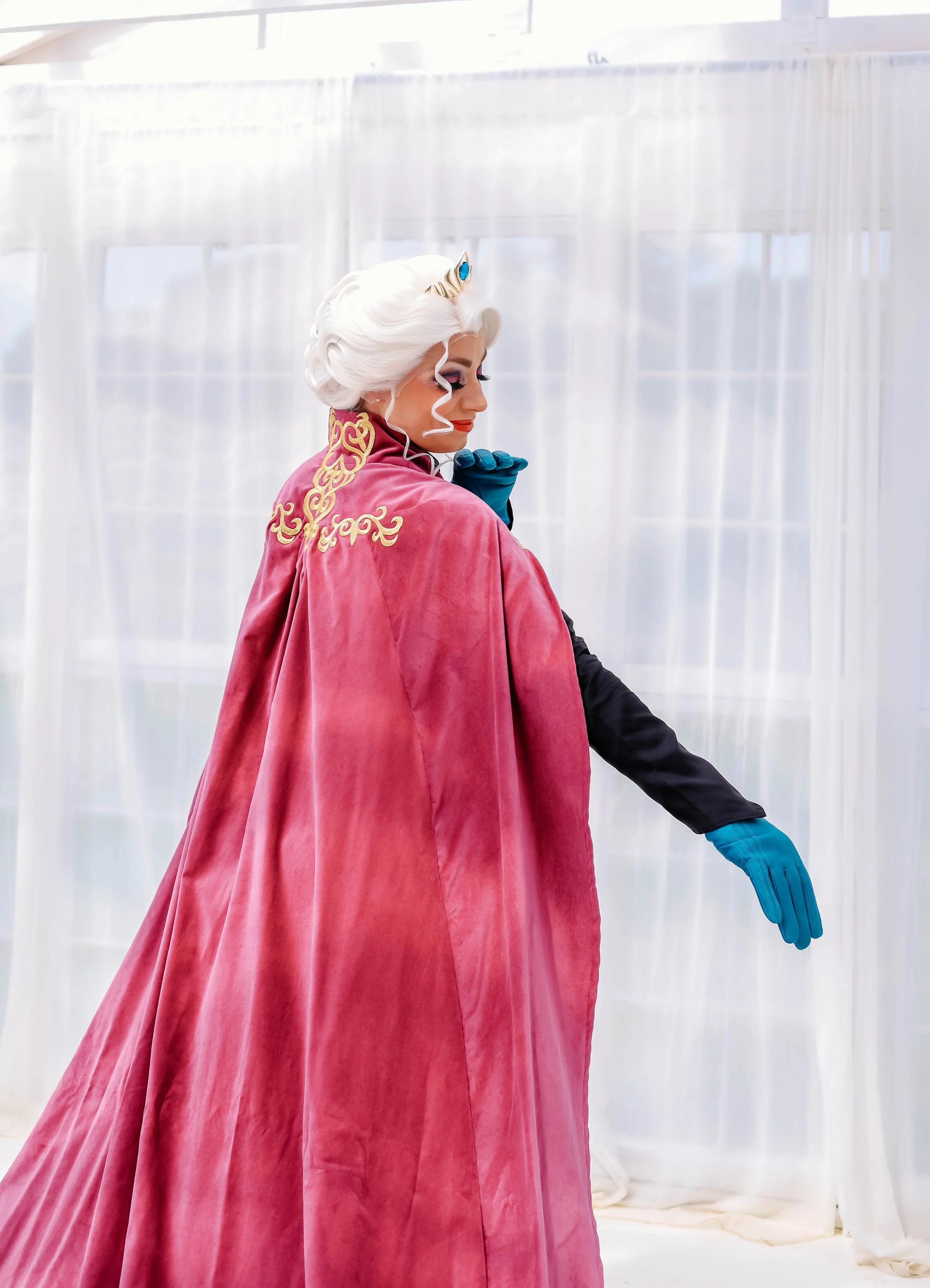 Person dressed as a princess or fairy tale character, wearing a long pink cape with gold embroidery, white curly hair, and a blue jewel tiara, standing in a bright, airy room with white curtains.