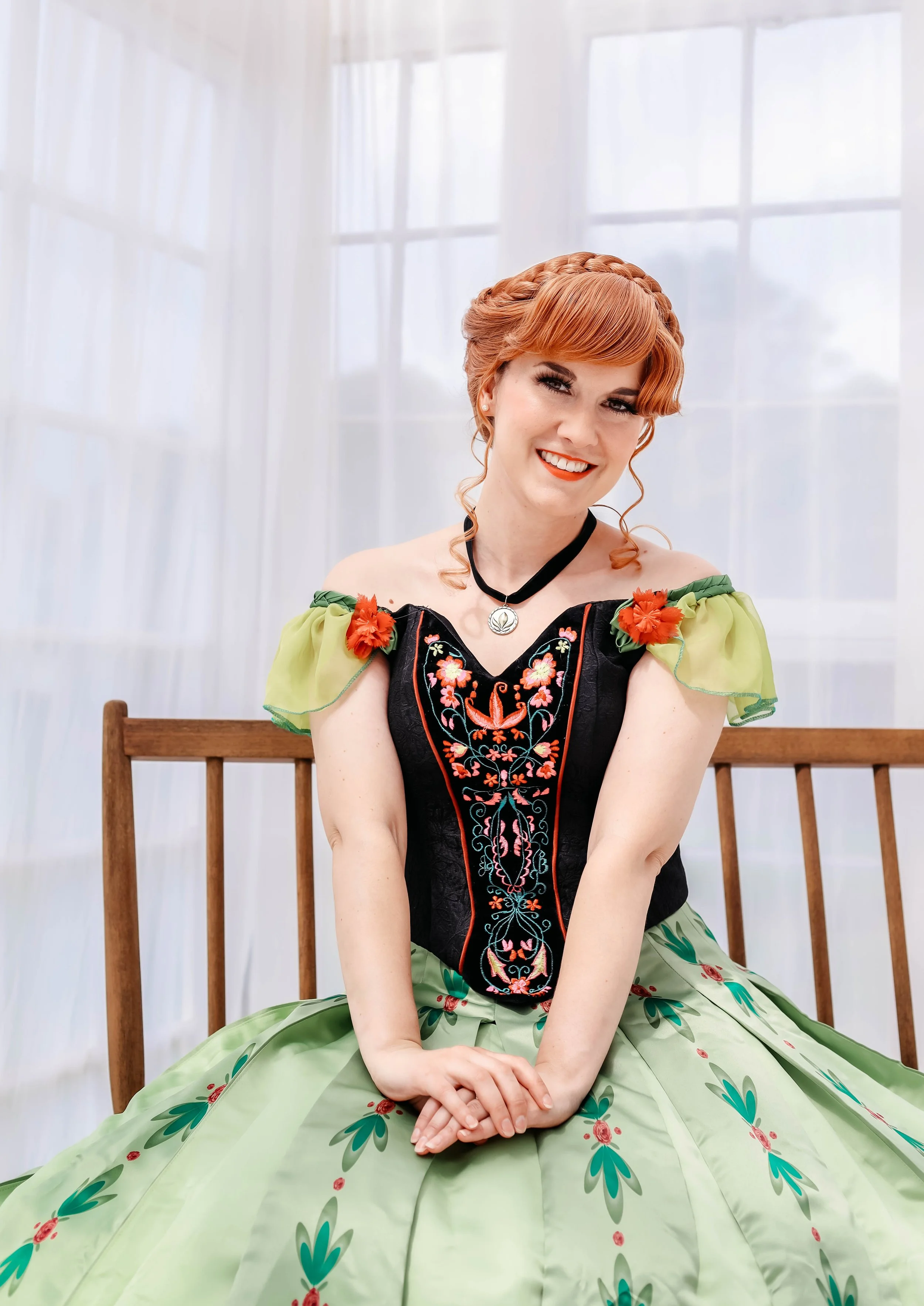A woman sitting on a wooden bench, wearing a colorful floral dress with green and black fabric and orange, pink, and green embroidery, in front of a sheer white curtain.