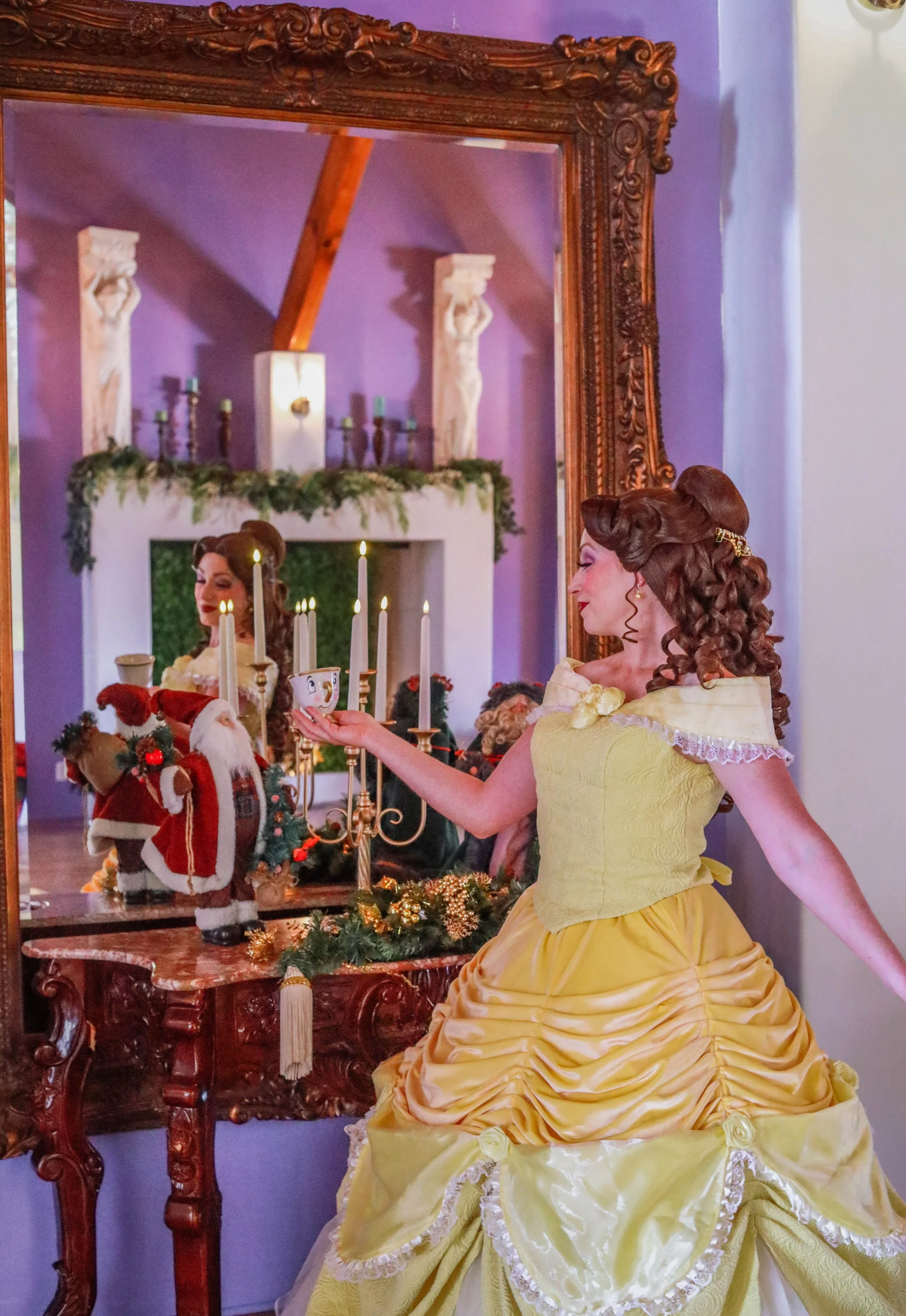 A woman dressed in a yellow ball gown, holding a small cup, standing in front of a large mirror with a reflection of a fireplace decorated with garland and candles, and some Christmas decorations including a Santa figurine and small Christmas tree.