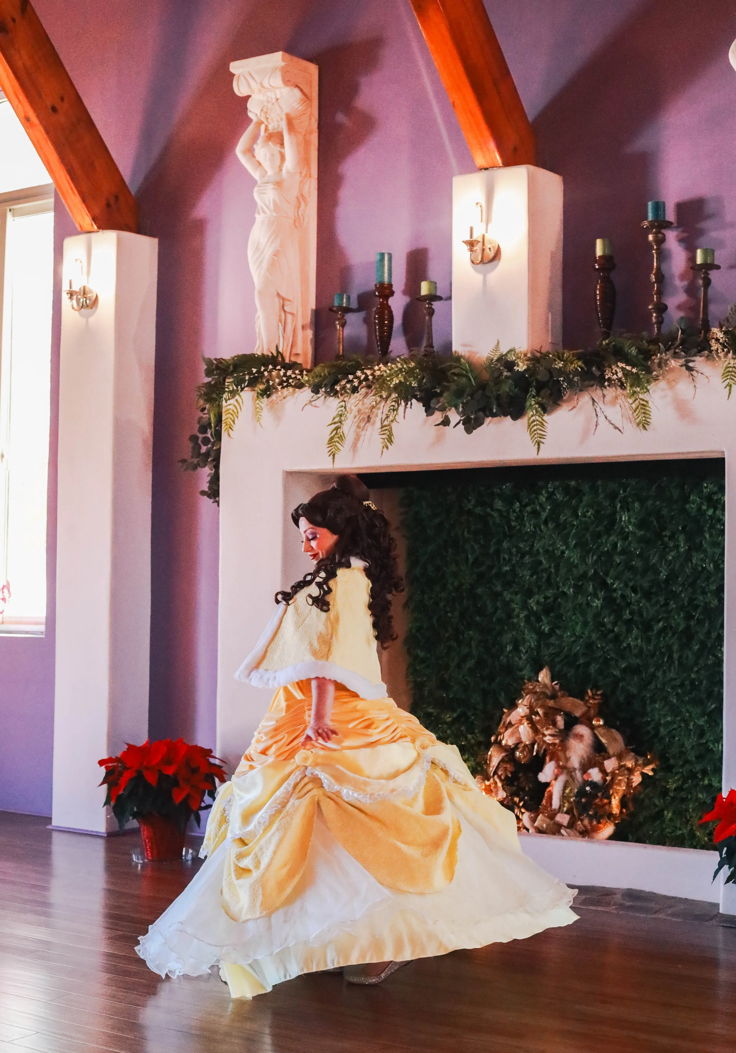 A woman in a yellow ball gown with a cream-colored cape, standing in a decorated room next to a fireplace with Christmas decorations and poinsettias.