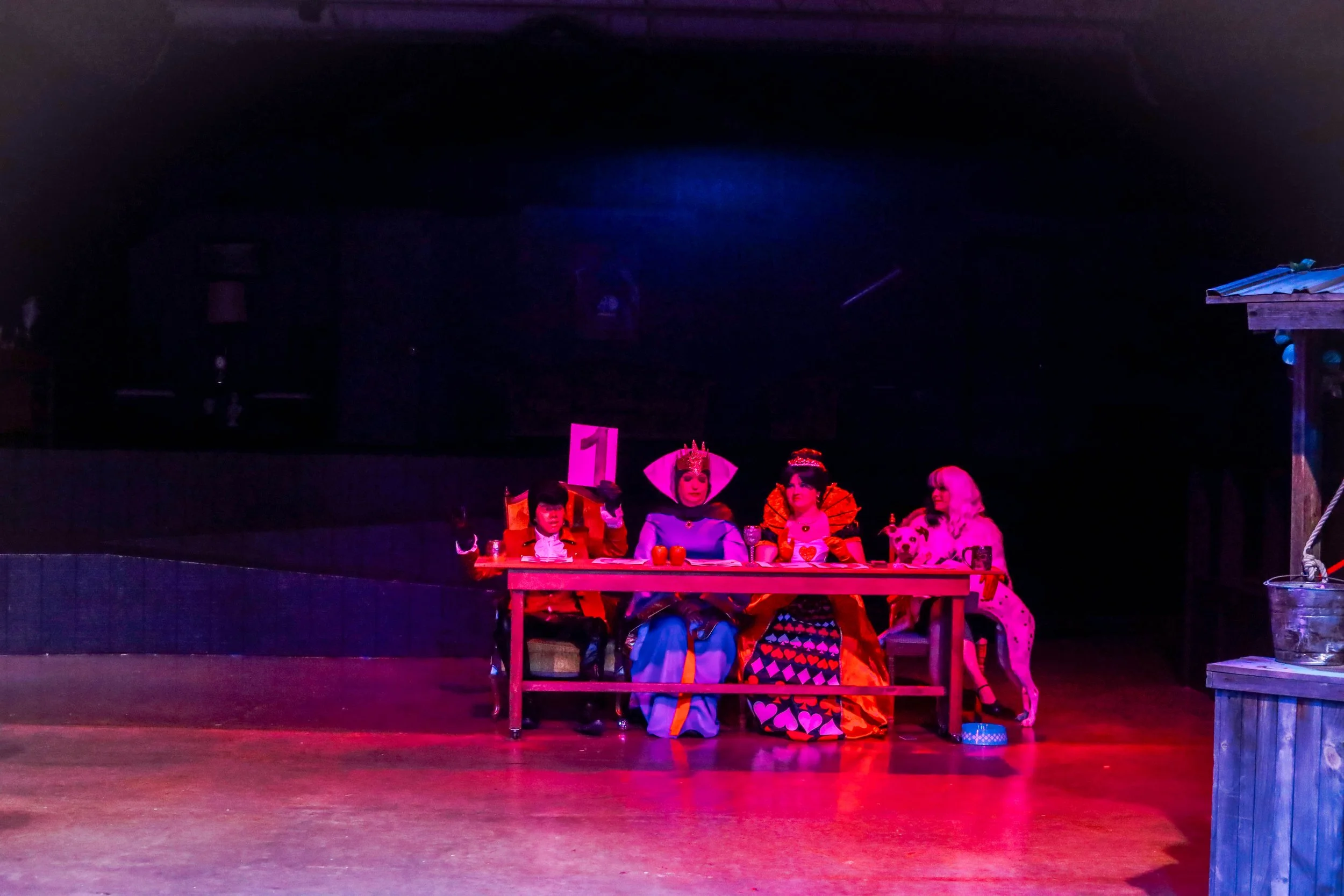 A theatrical stage scene with four characters in colorful costumes sitting at a table, with a pink number 1 sign behind them, in a darkened theater with a wooden floor and minimal background elements.