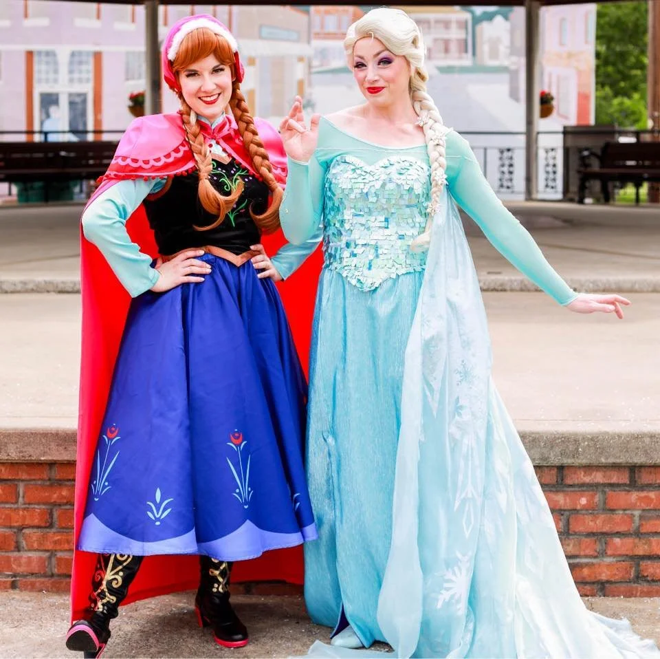 Two women dressed as sister princesses, posing outdoors with buildings and benches in the background.