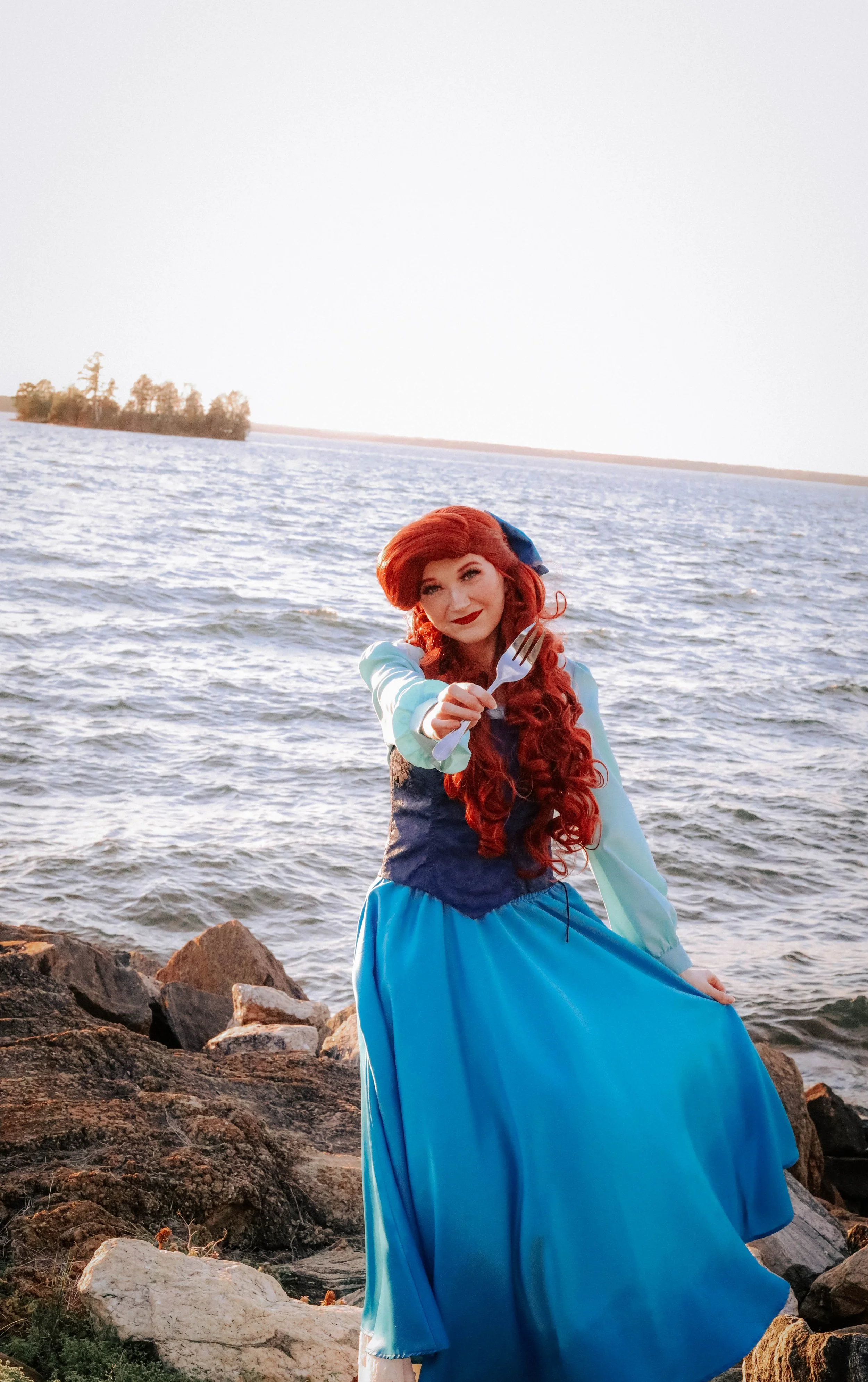 A woman dressed as a mermaid, with long red curly hair, wearing a blue dress and holding a plastic fork, standing on rocks by a body of water during sunset.