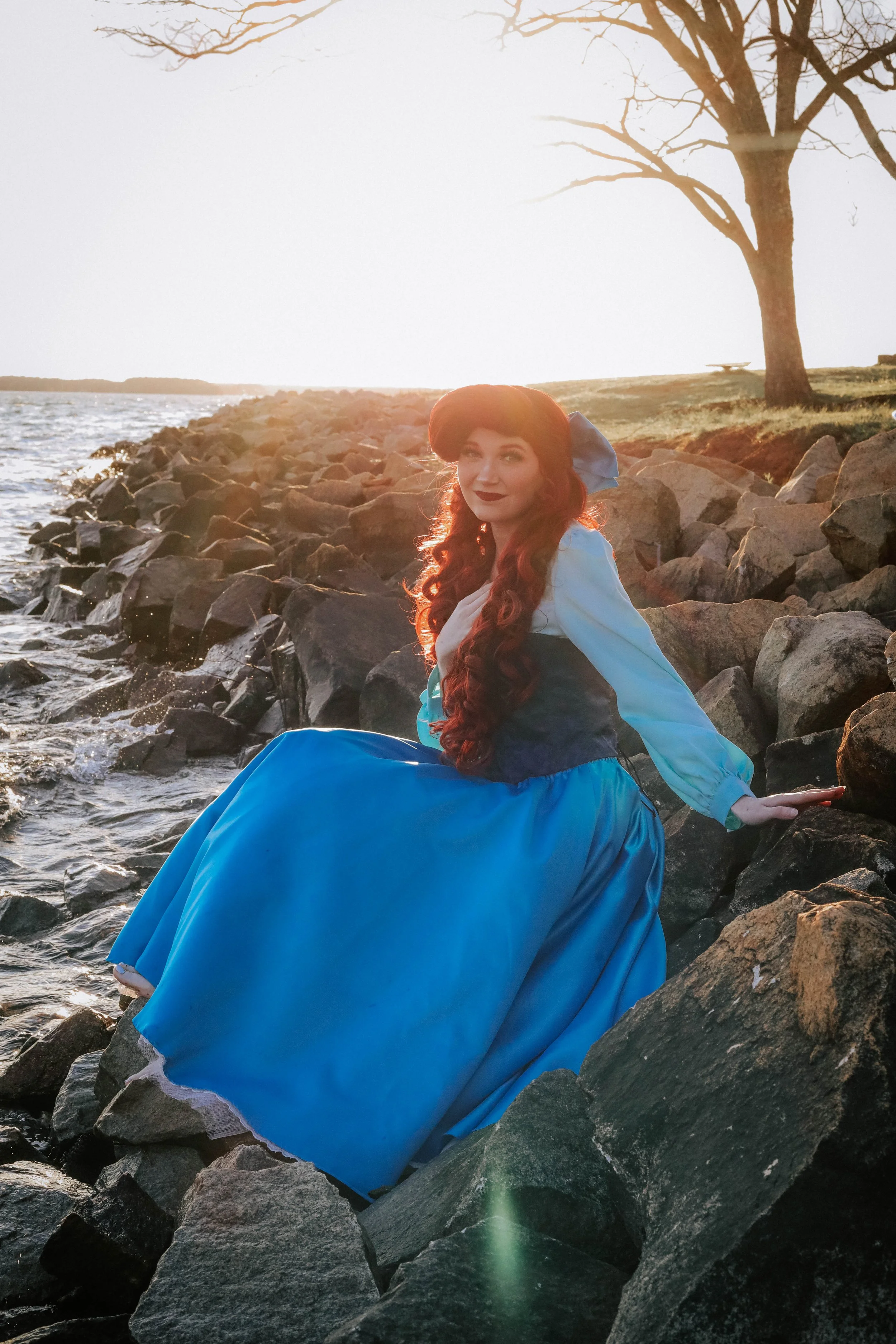 A woman dressed in a blue and white historical costume sitting on rocks by the water at sunset, with leafless trees in the background.