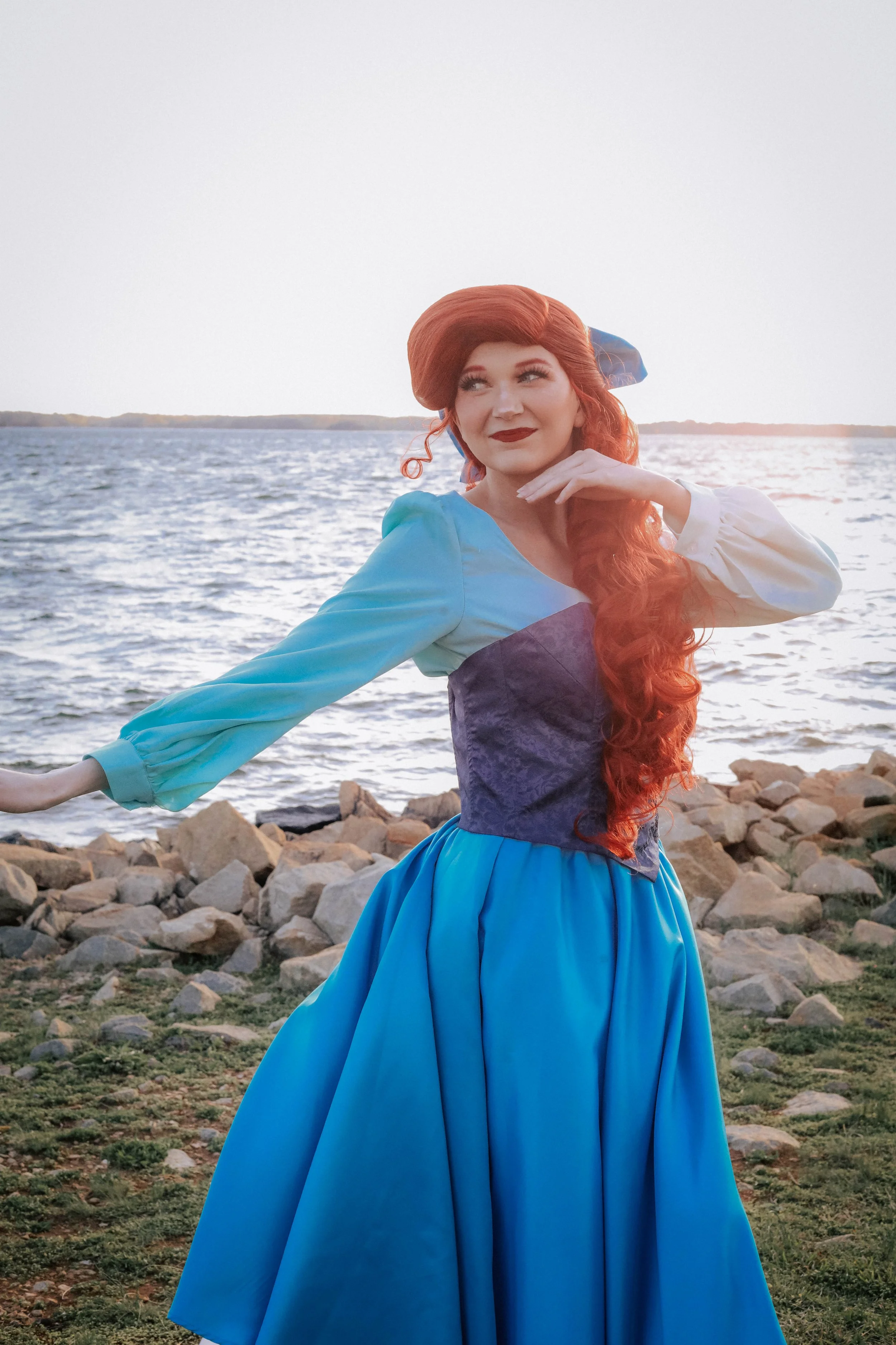 A woman with long red hair, wearing a blue dress with puffy sleeves, stands near a rocky shoreline with water in the background, during sunset.