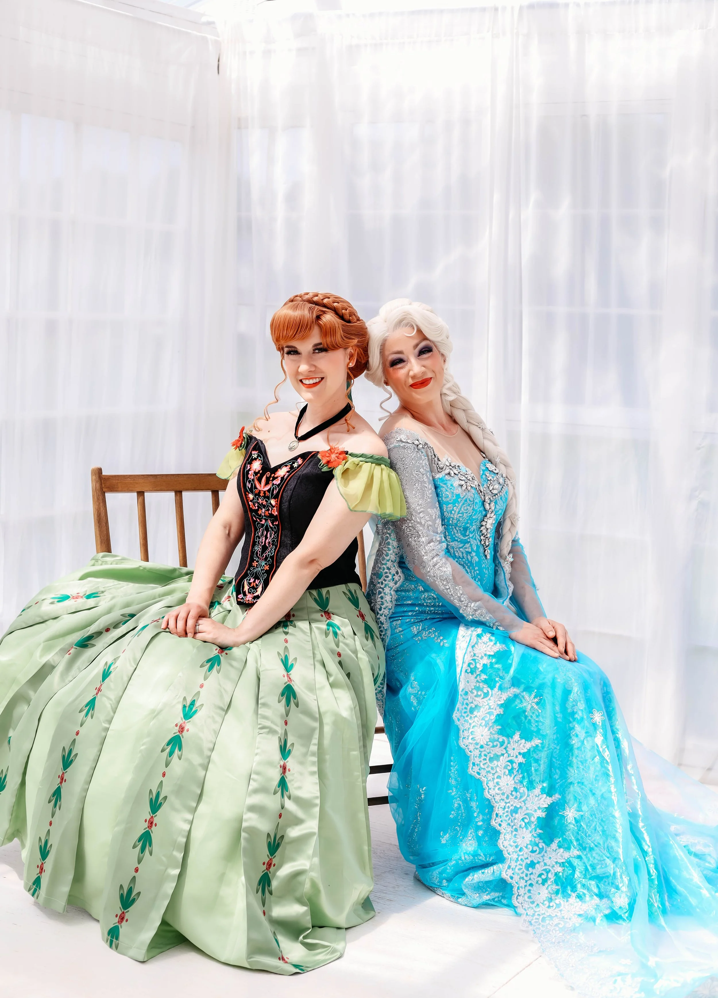 Two women dressed as princesses sitting side by side on a wooden bench, in front of sheer white curtains.