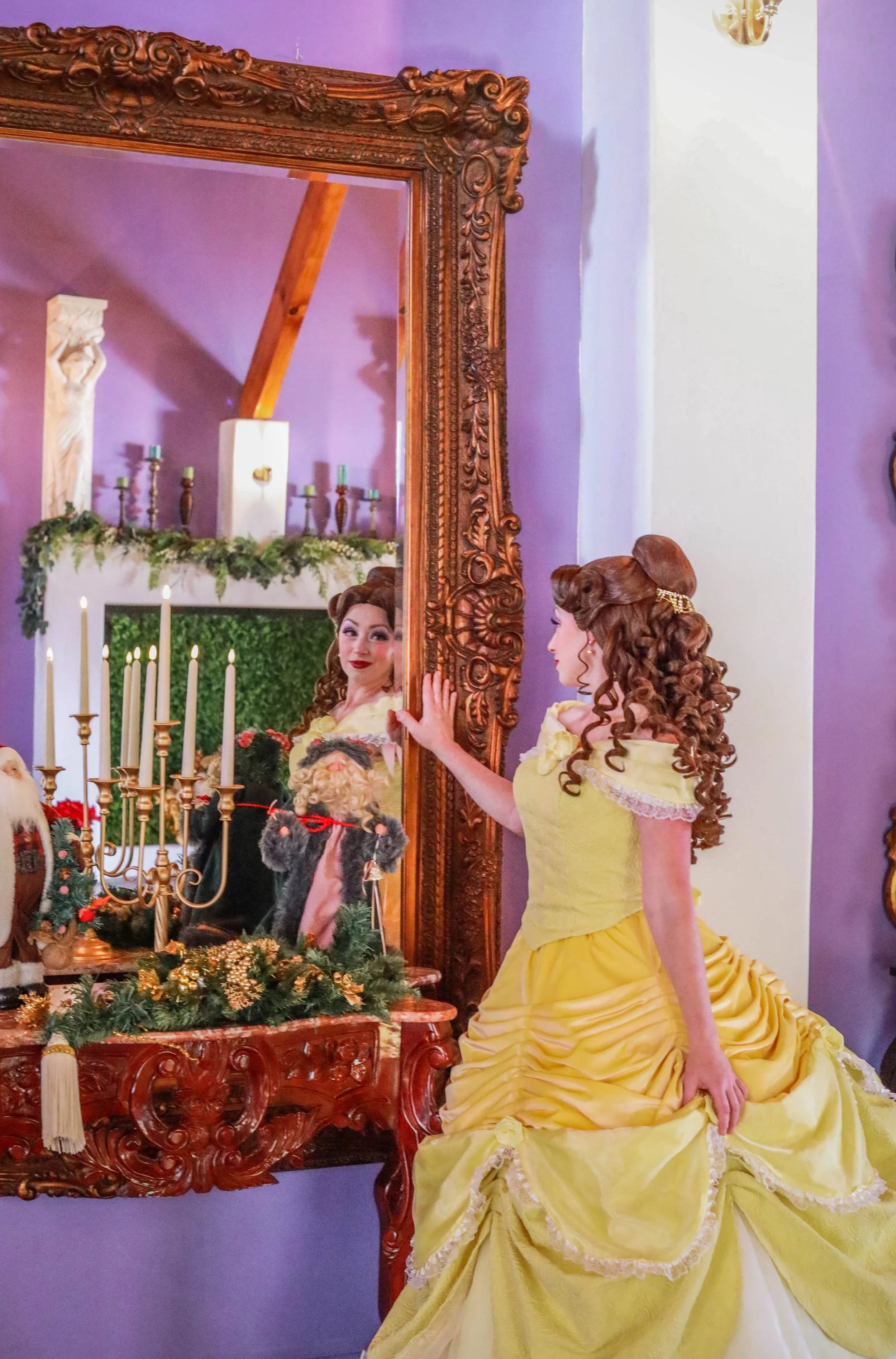 A woman dressed as a princess in a yellow ball gown standing in a decorated room, looking at her reflection in a mirror.
