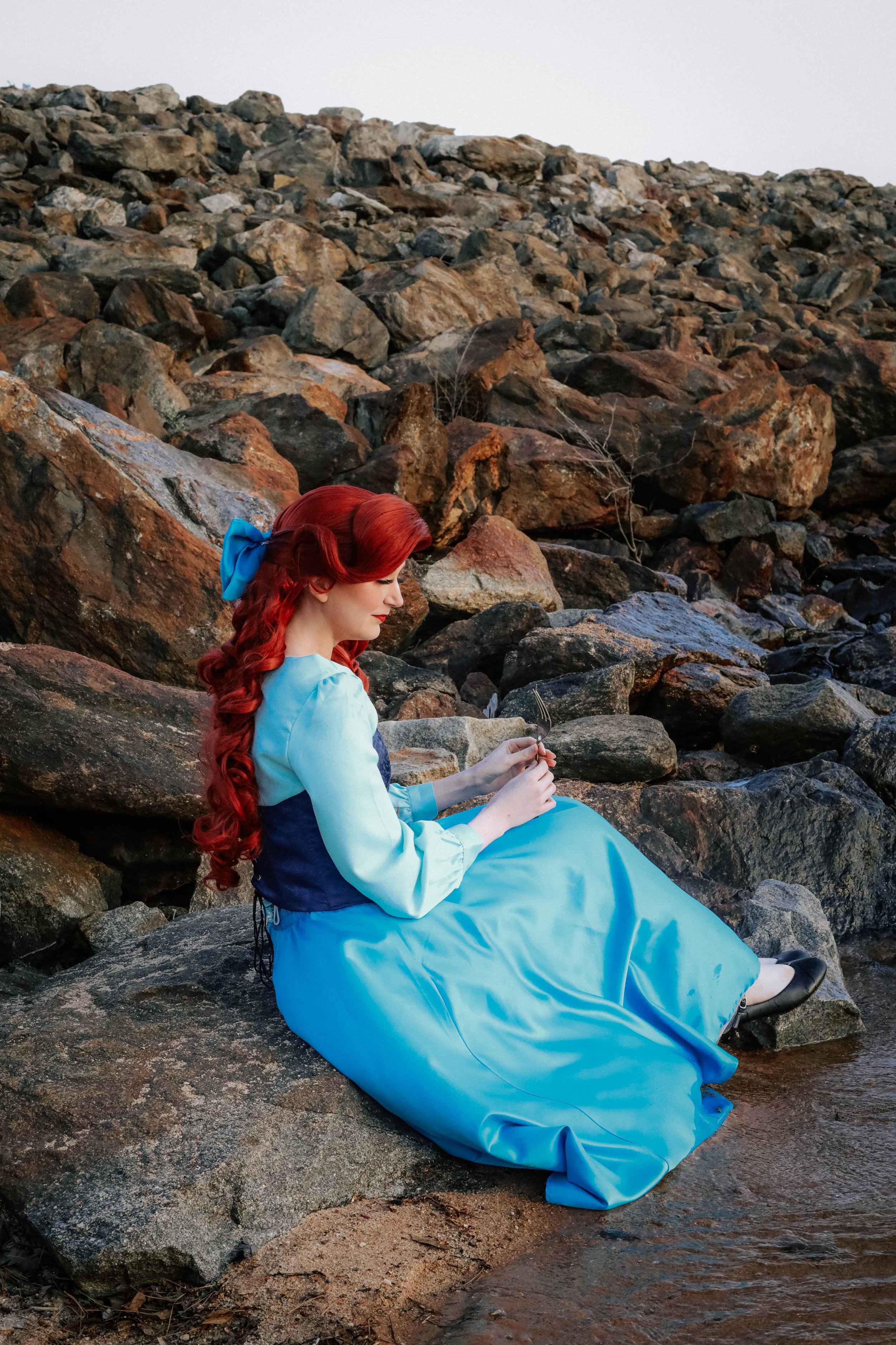 A woman with long red hair, dressed in a blue gown, sitting on a rock beside a stream, holding a small fork and a plate, with a rocky hillside in the background.