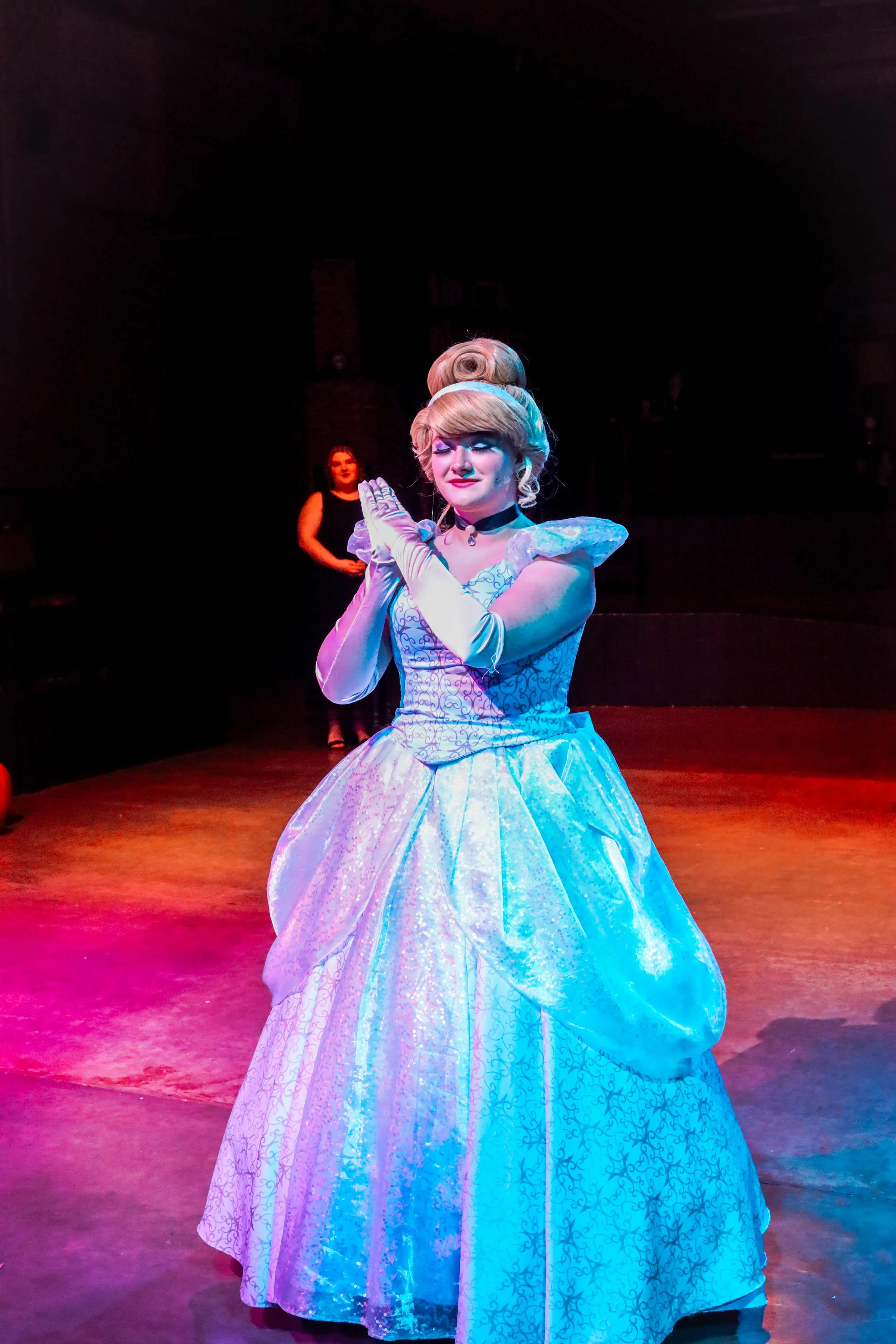 A woman dressed as a princess or fairy tale character in a light blue gown, standing with hands clasped together under theatrical stage lighting, with another woman in the background on a dimly lit stage.