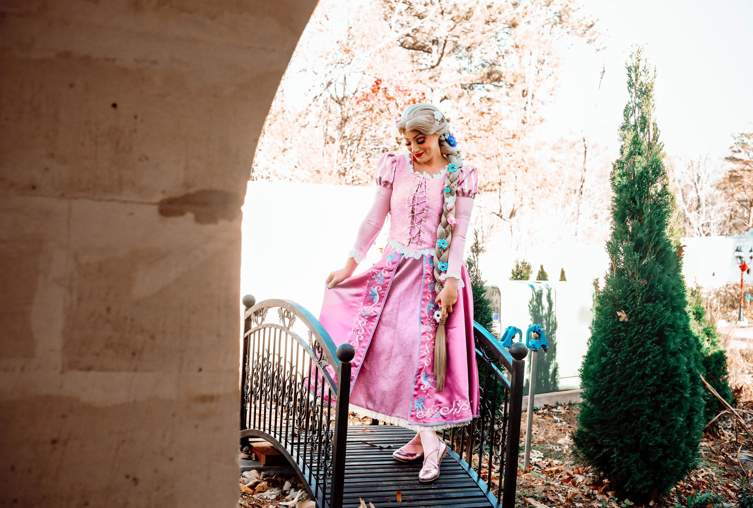 A woman dressed as a princess in a pink gown with embroidered details, standing on a small decorative bridge outdoors with trees and a white fence in the background.