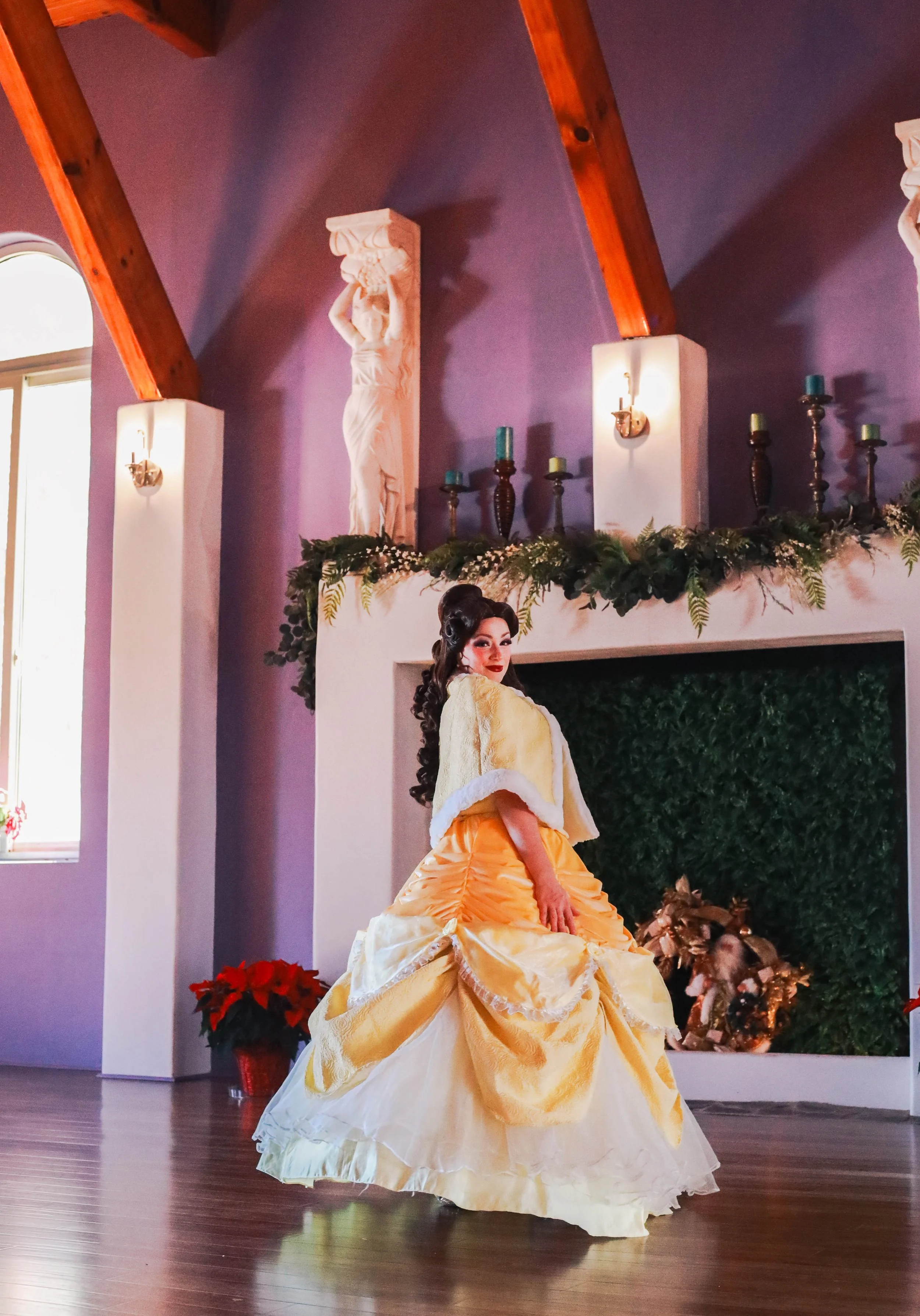 A woman in a yellow ball gown with ruffles and a white cape, standing in a decorated room with purple walls, a fireplace, and holiday decorations.