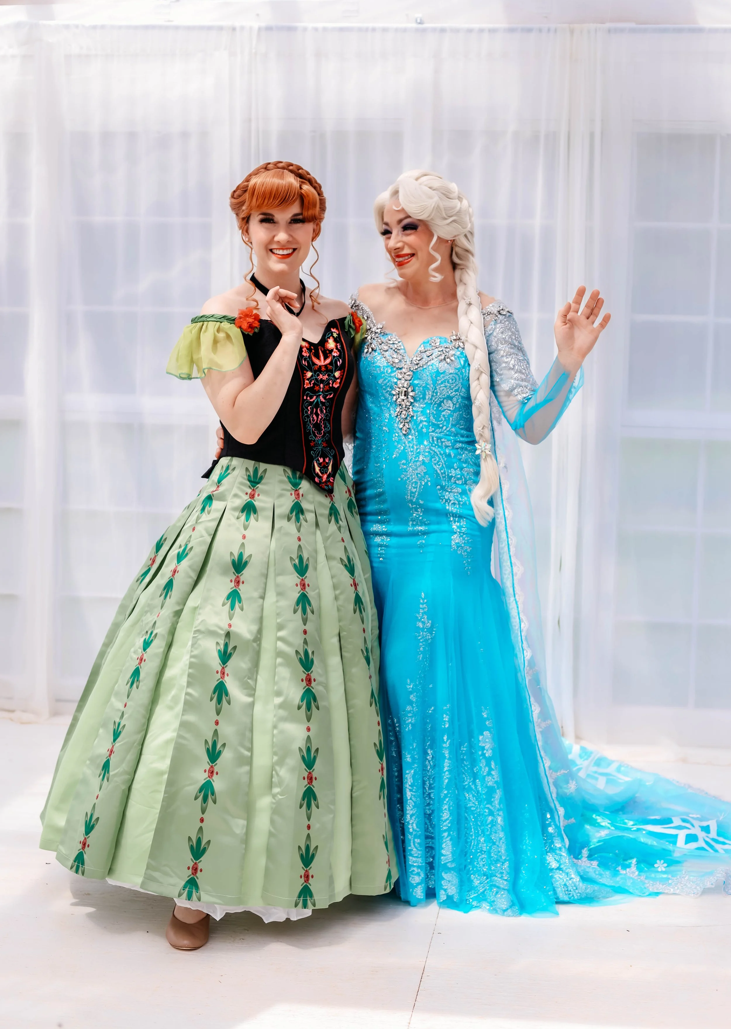Two women dressed as princesses, standing together smiling, with one wearing a green and black dress with floral patterns and one in a blue icy gown with lace details.