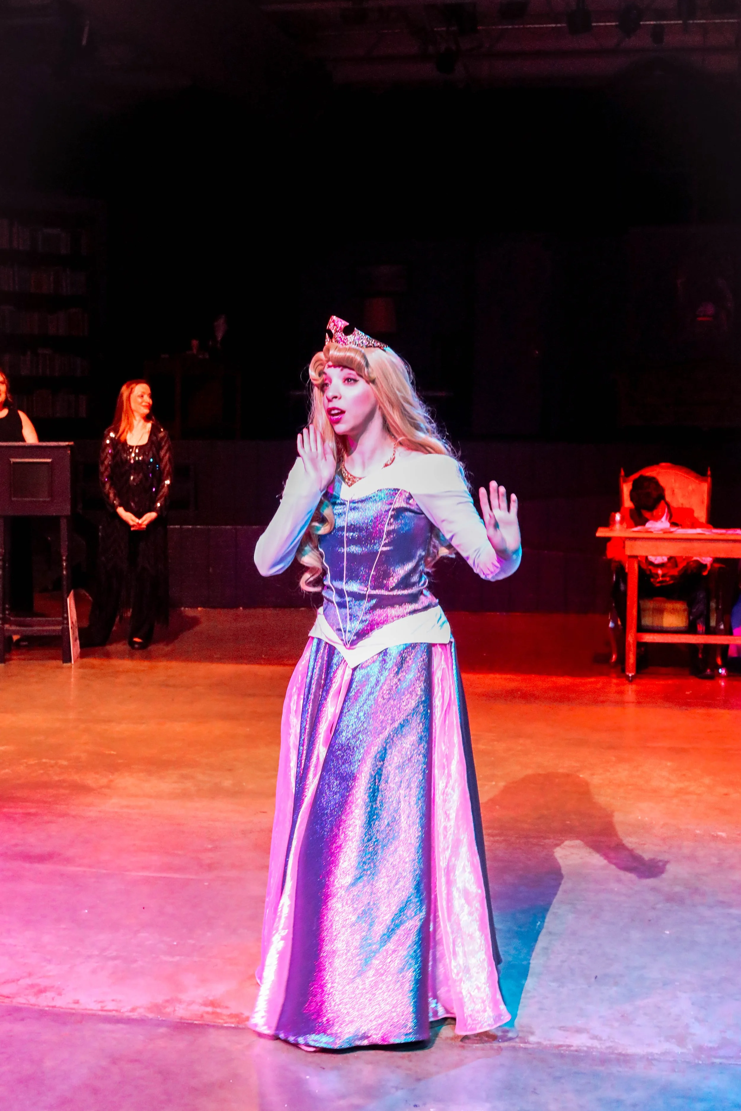 Actress dressed as a princess in a shimmering gown and crown, performing on stage with other cast members in the background.