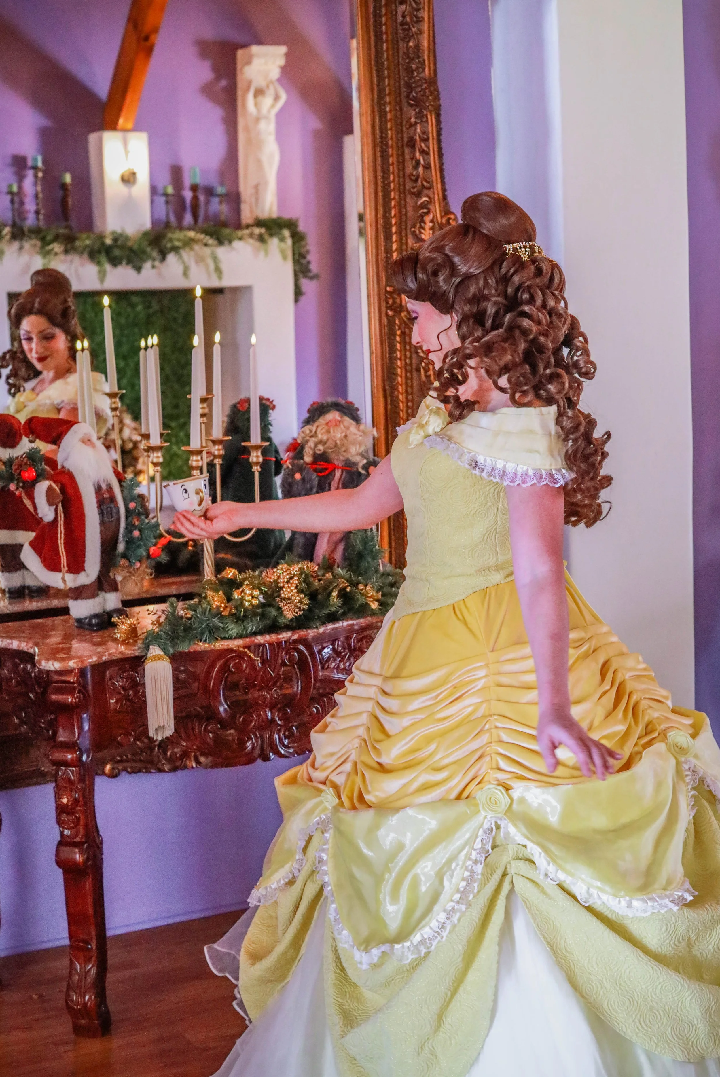 A woman dressed as a princess wearing a yellow gown with ruffles, stands in front of a mirror in a decorated room with Christmas decorations, candles, and plush Santa and reindeer figures.
