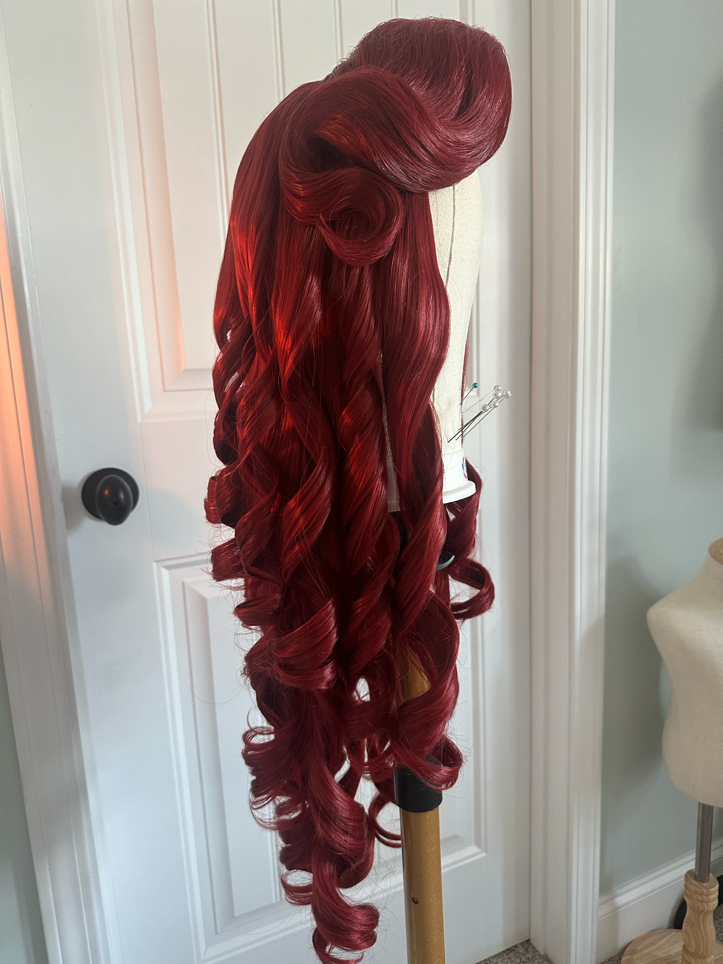 A red, curly wig styled in vintage curls displayed on a mannequin head, mounted on a stand in a room with white door and light-colored walls.