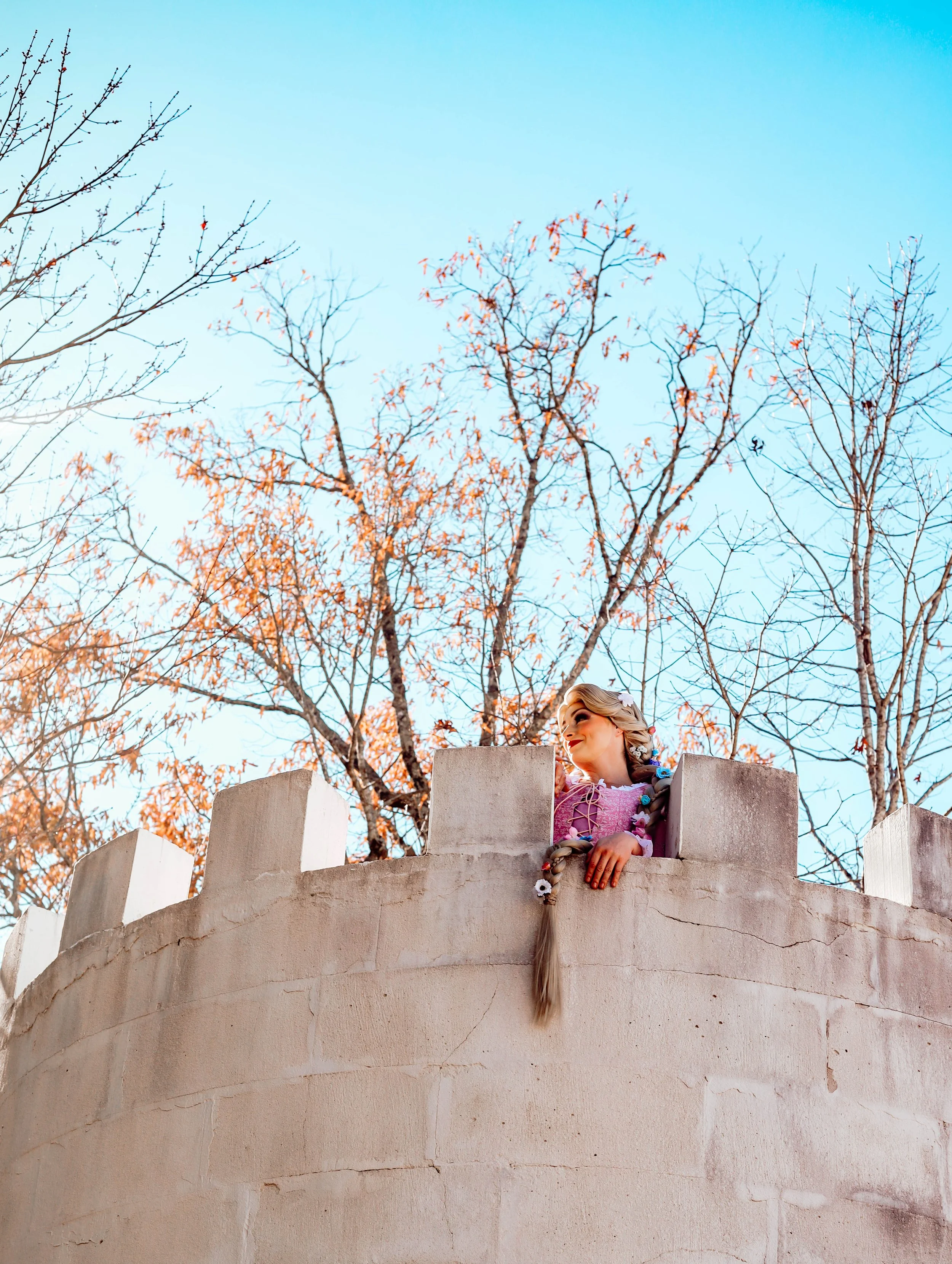A woman with long braided hair leaning over the top of a stone castle wall, smiling, with leafless trees and a clear blue sky in the background.