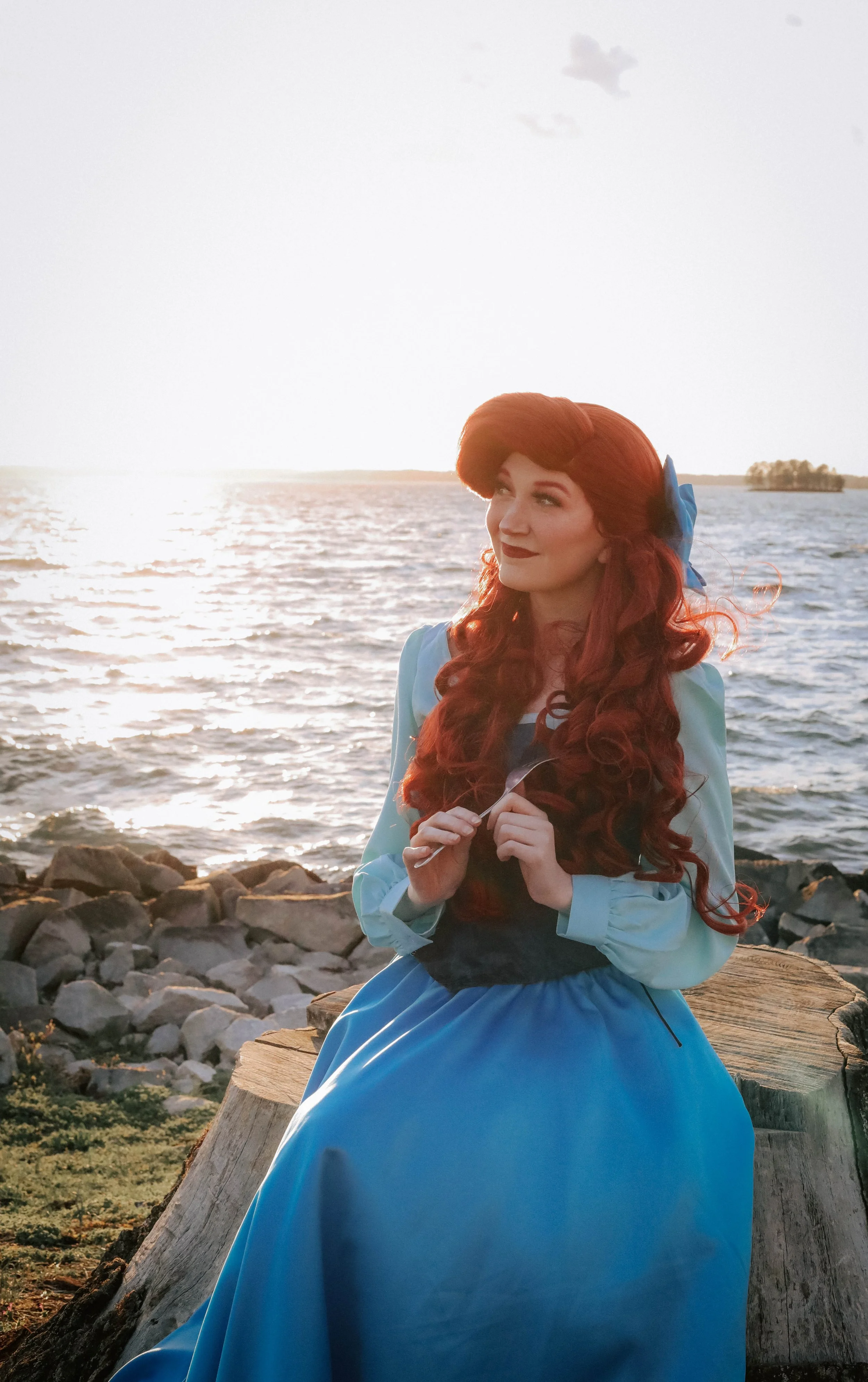 A woman dressed as a Mermaid, sitting on a tree stump near a body of water at sunset, holding a fork, with long red hair and a blue bow in her hair.