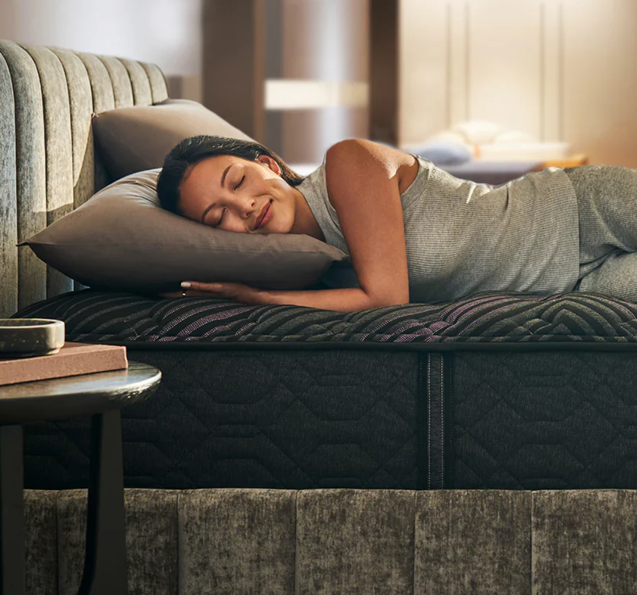 Woman lying on her side in bed, smiling, resting her head on a gray pillow, in a bedroom with warm lighting.