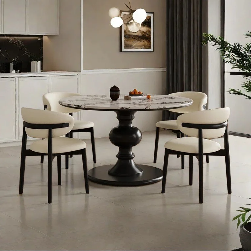 Modern dining room with a round marble table, four cream-colored chairs with black legs, a chandelier with globes, a painting on the beige wall, and a large window with dark curtains.