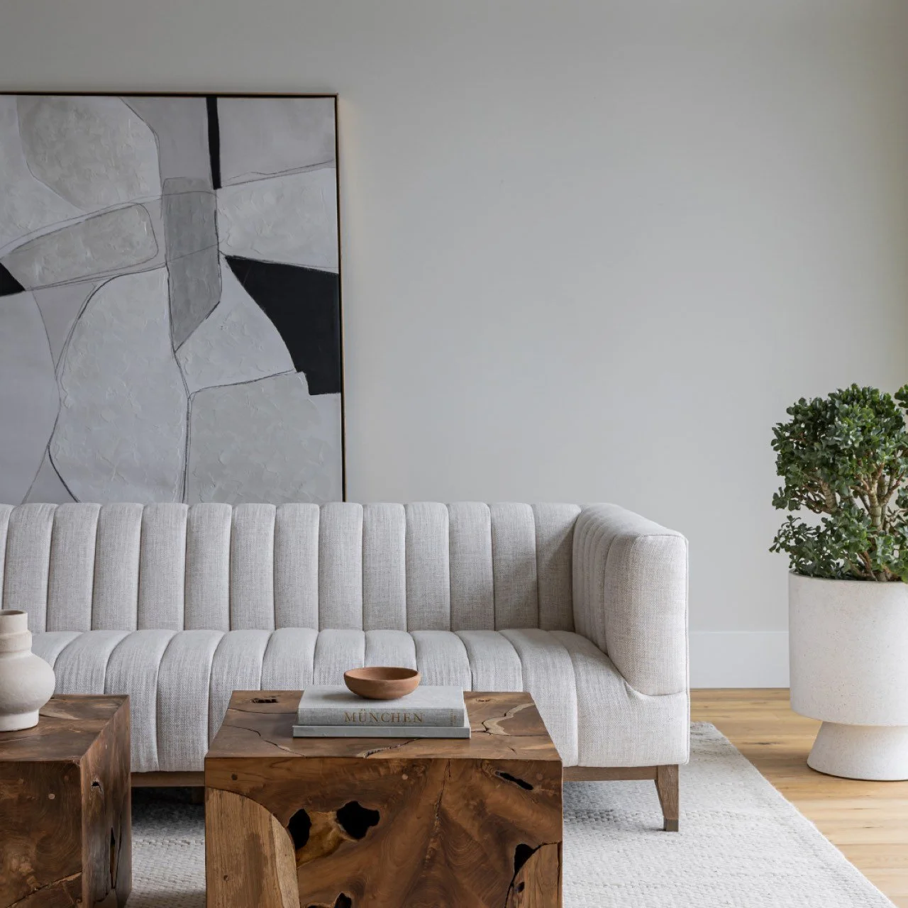 A modern living room with a light-colored, upholstered sofa, abstract black and white artwork on the wall, a wooden coffee table with a book and a small bowl, a white vase, and a large potted plant.