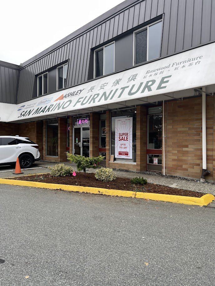Exterior of a furniture store called San Marino Furniture with a promotional sign for a super sale offering up to 70% off, parked white car, and small landscaped area with plants and a tree in front.