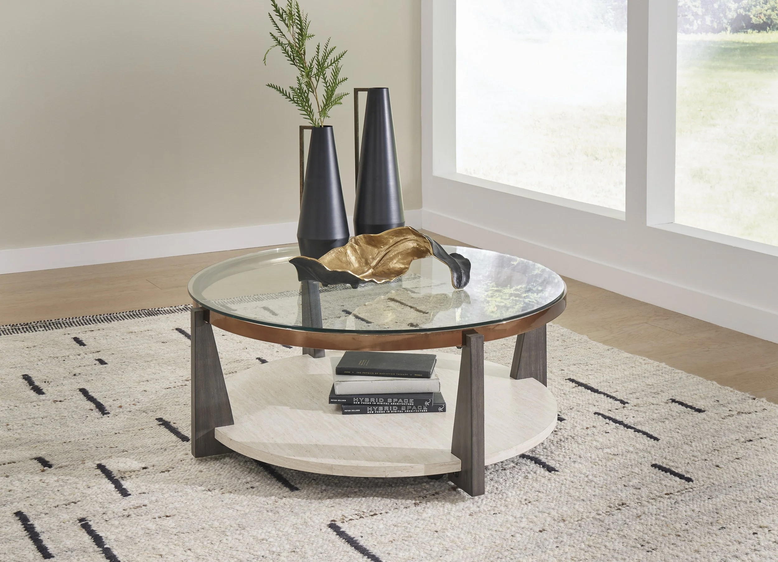 Modern living room corner with a round glass coffee table, two tall black vases with greenery, and a decorative black and gold sculpture on the table, placed on a neutral area rug.