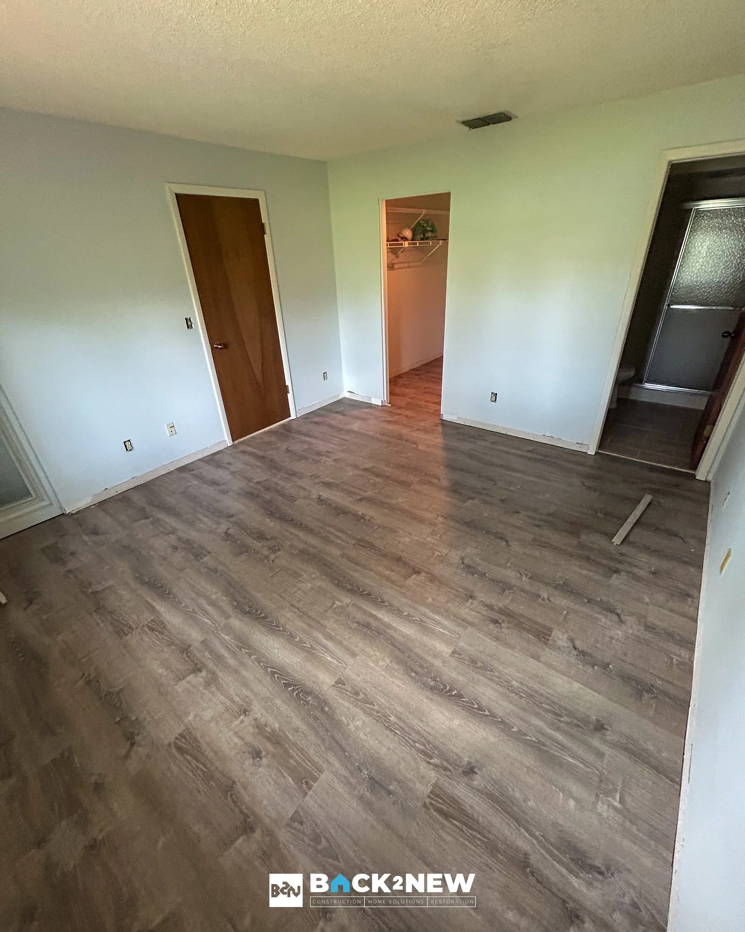 Empty room with light-colored walls, wood flooring, a closed wooden door, and an open closet with shelves, part of a bathroom with a shower, and a vent on the ceiling.