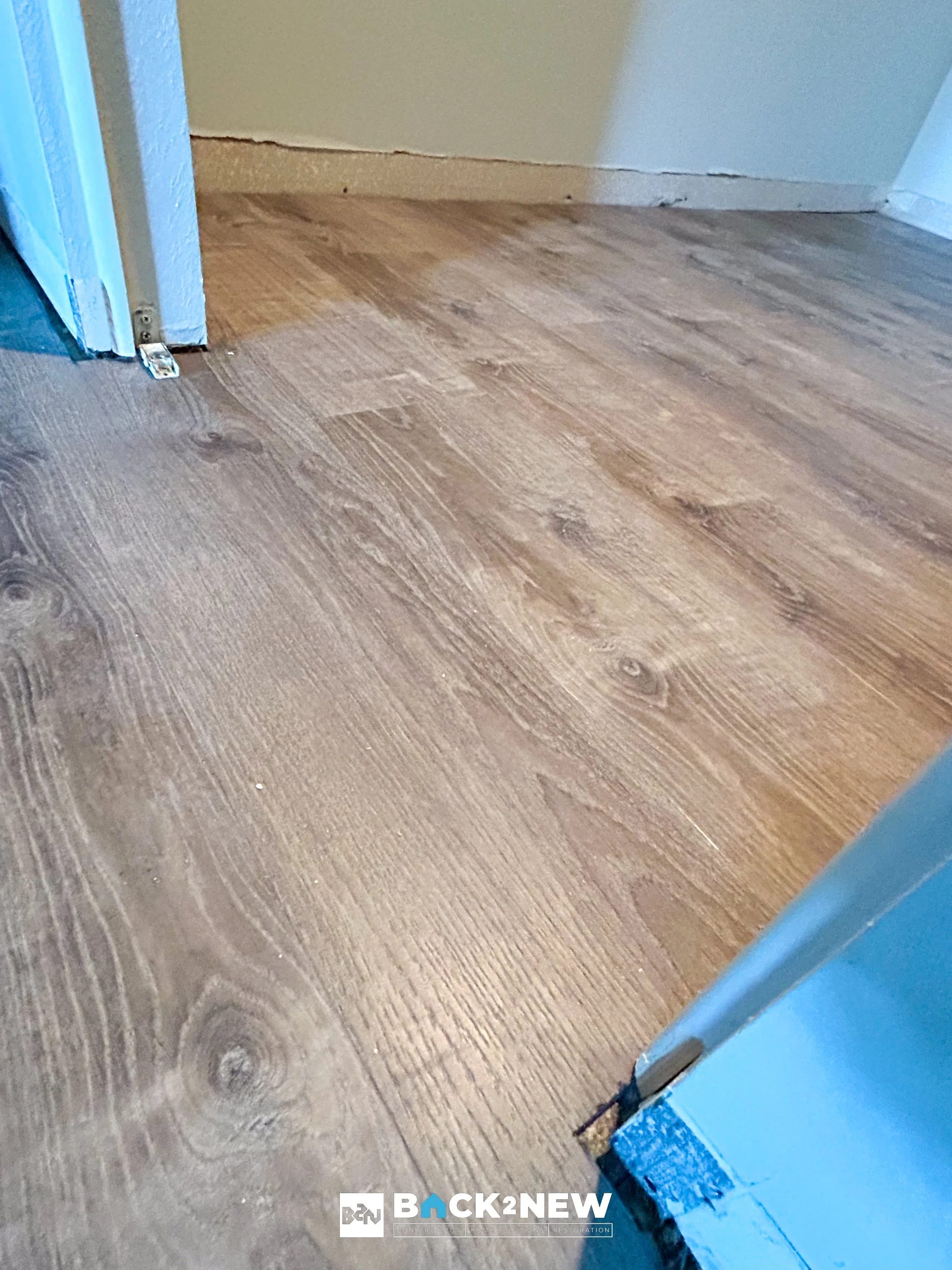 Wooden floor in a room with a beige wall and blue door frame, showing a transition strip or gap at the doorway.