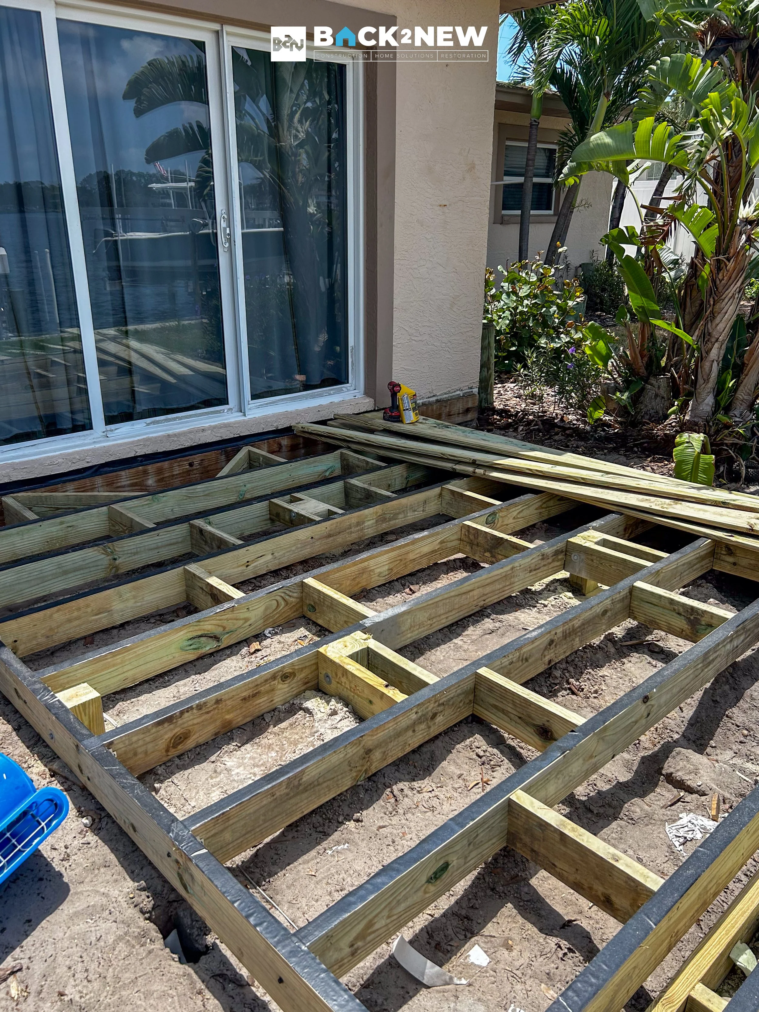 Wooden framework construction for a deck outside a house with sliding glass door, tools, and tropical plants nearby.