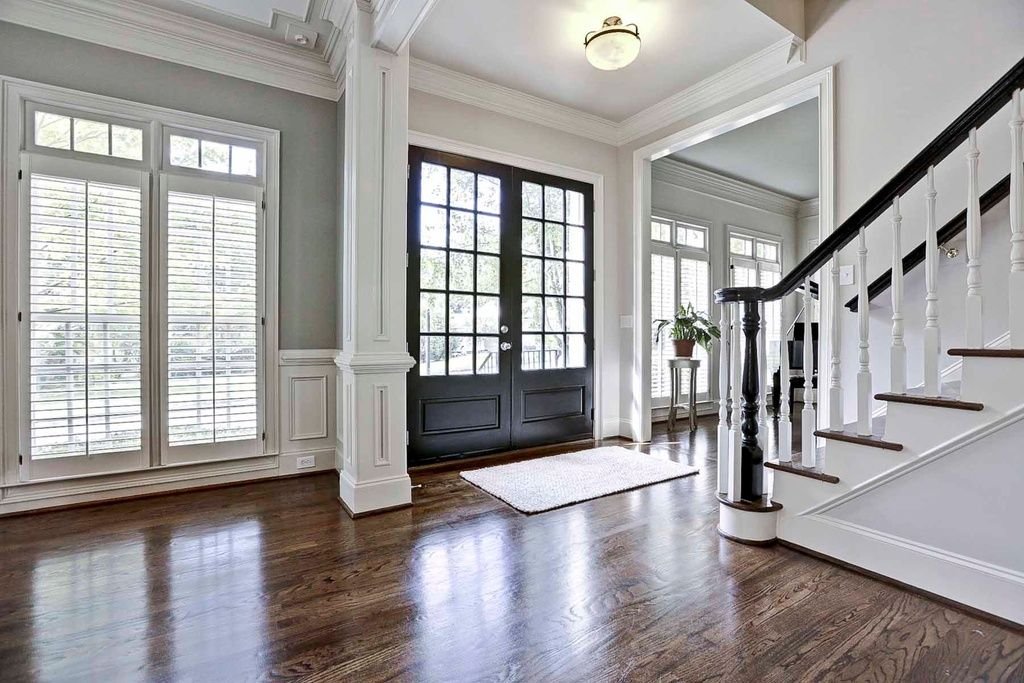 Elegant entryway of a house with a black front door with glass panes, large windows with white blinds, a small side table with a potted plant, hardwood floors, and staircase with white spindles and dark handrail.