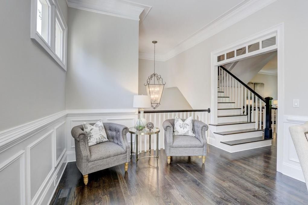 Stunning living room corner with two gray upholstered armchairs, a small round side table with a lamp, and a chandelier overhead. Expert white wainscoting and staircase in background.