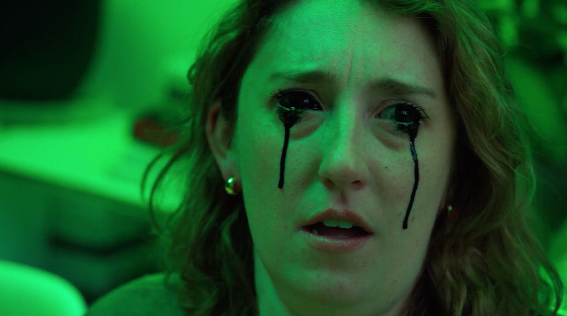 A young woman with curly hair and earrings, face illuminated by green light, with tears black makeup running down her cheeks.