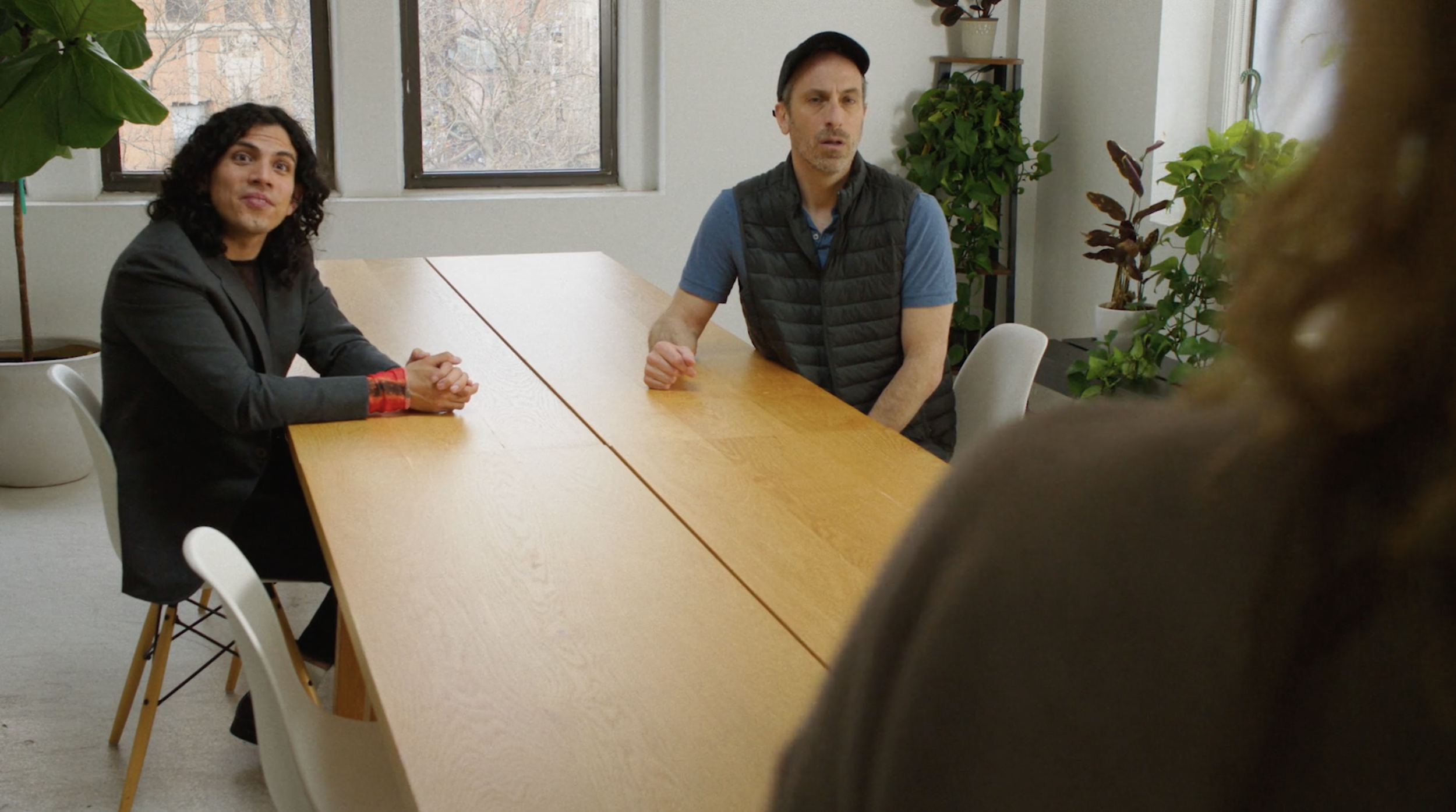 Two people sitting at a wooden table in a room with large windows and potted plants, watching a pitch.