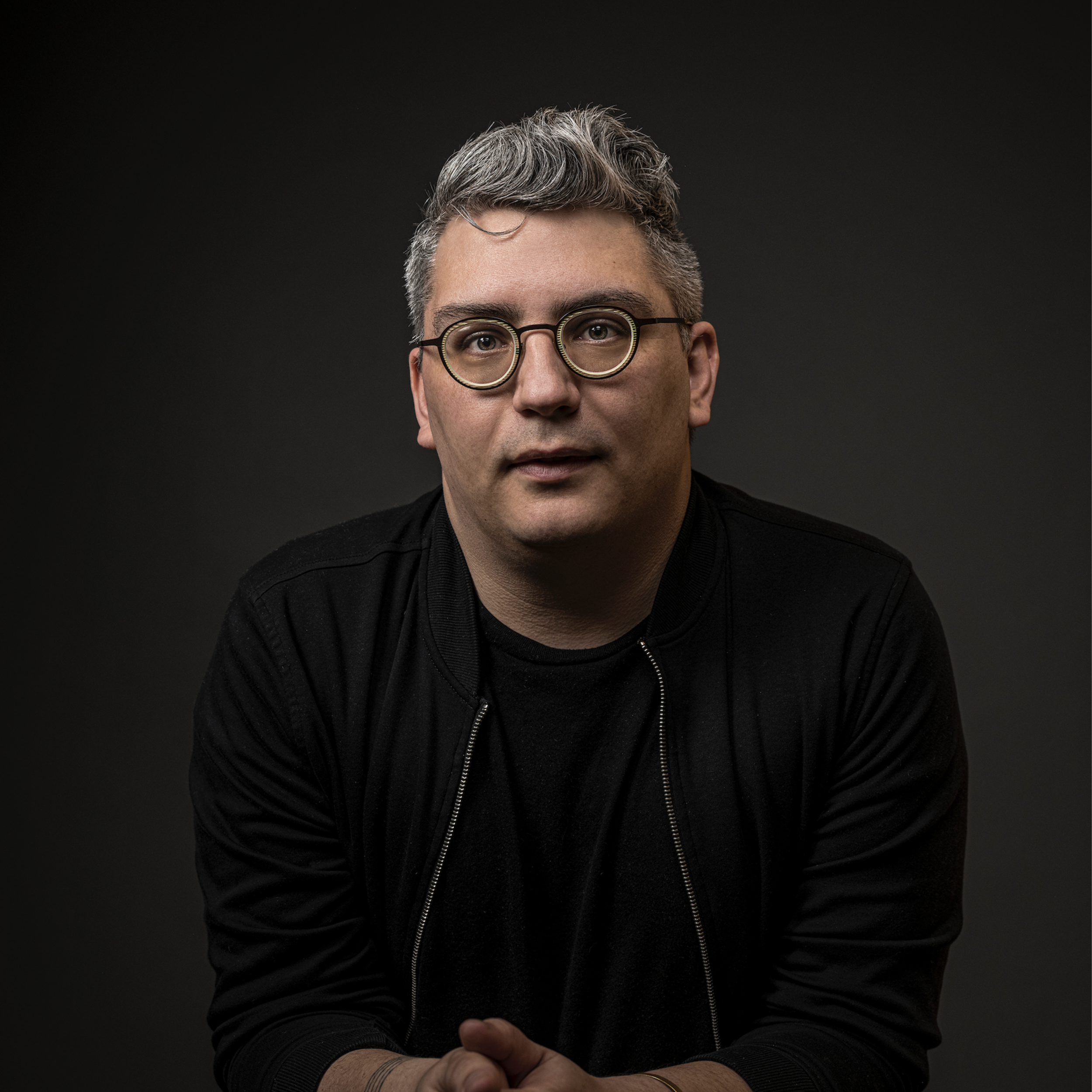 A man with grayish hair wearing black-rimmed glasses and a black jacket, looking at the camera against a dark background.
