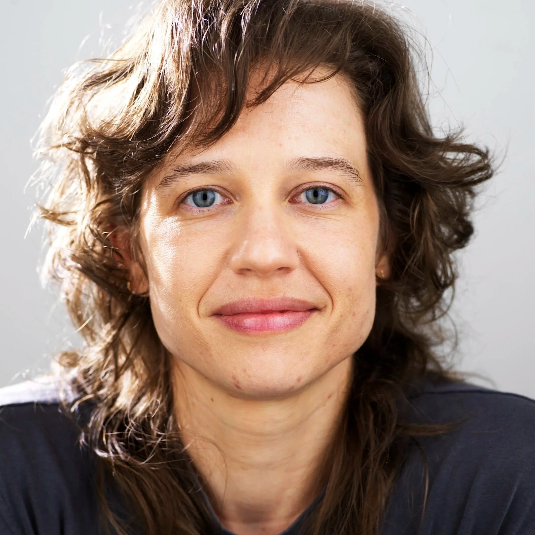 Close-up of a persons face with gray-blue eyes, brown curly hair, and a slight smile. They are wearing a dark top.