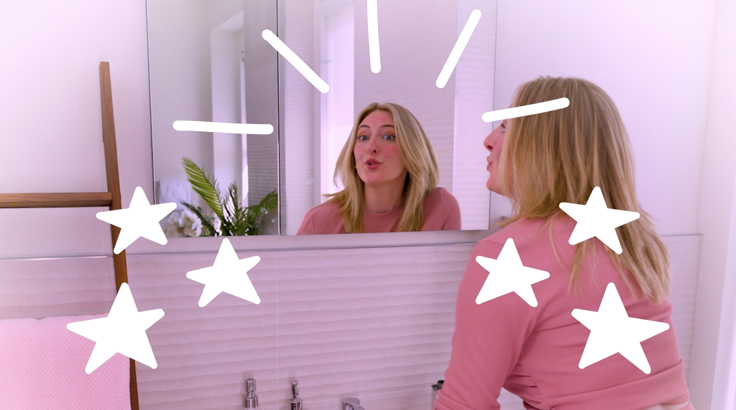 Woman with blonde hair looking at herself in a mirror in a pink room decorated with white star illustrations.