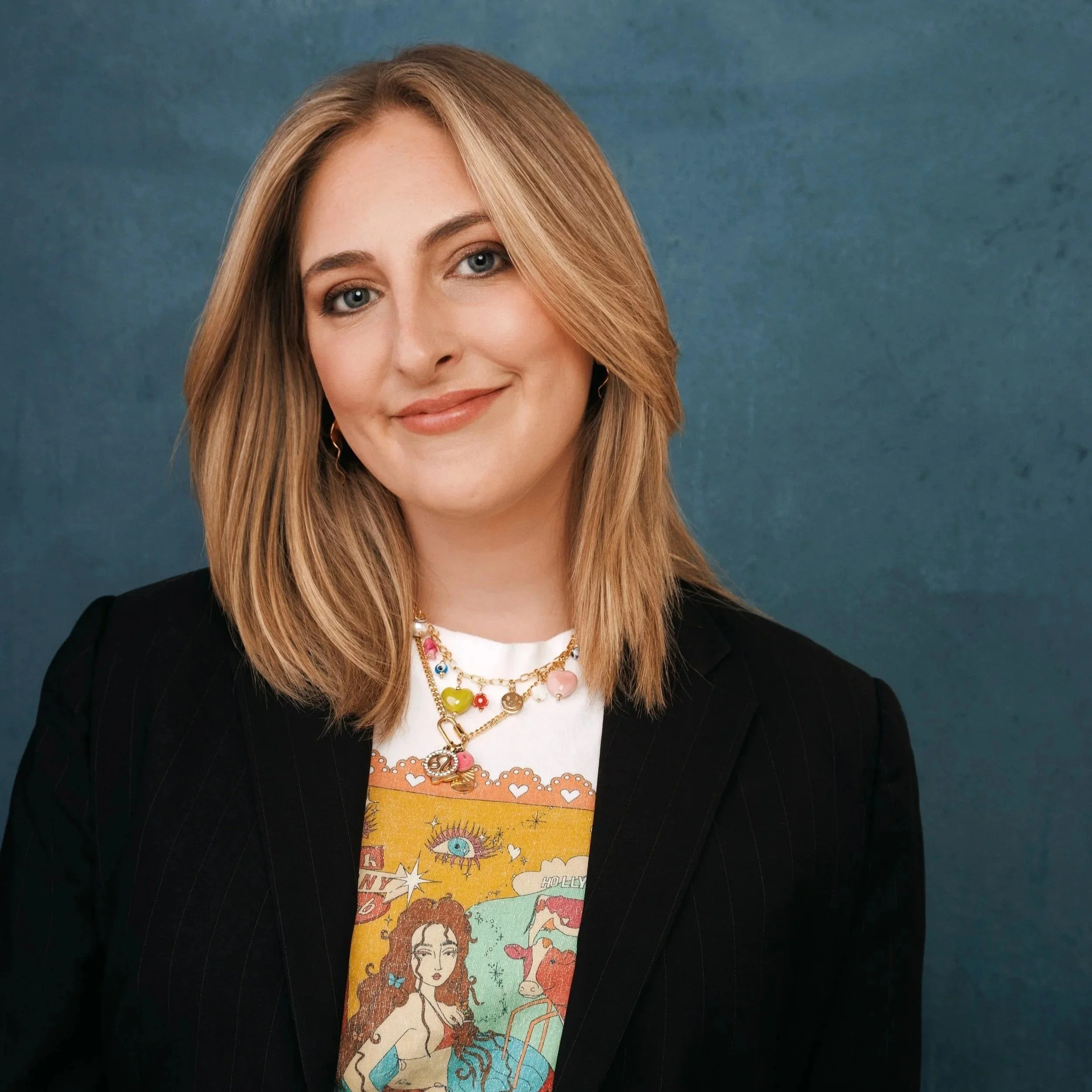 Close-up of a woman with long, blonde hair styled in loose waves, smiling softly, wearing gold hoop earrings, layered necklaces, a white top with a colorful illustrated graphic of a woman with long hair, and a black blazer, against a textured blue background.