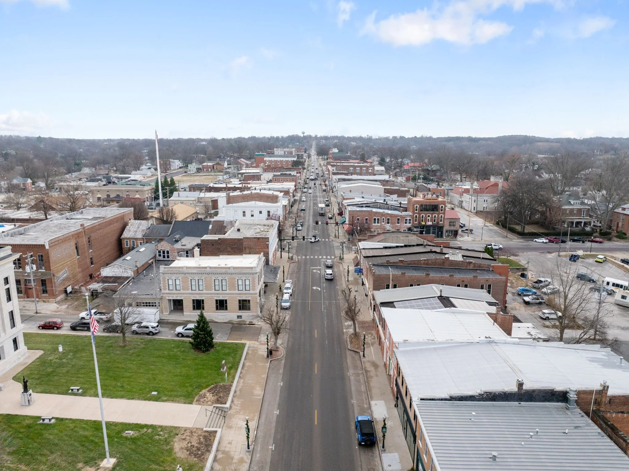 Aerial view of a small town with a main street running through the center, lined with brick and stone buildings, cars parked along the street and on side streets, and a small park with a flagpole and statues in the foreground. The town is surrounded 