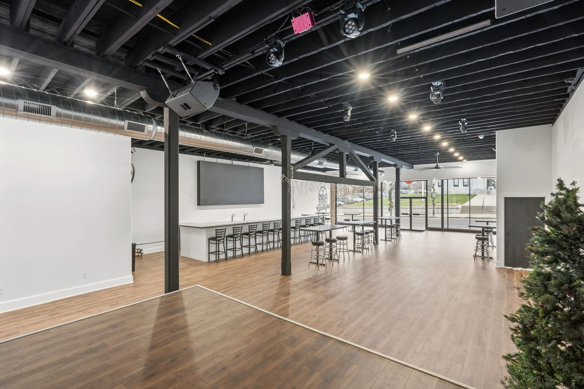 Empty indoor event space with wooden flooring, white walls, black ceiling, large windows, and minimal decor including a small Christmas tree in the foreground.