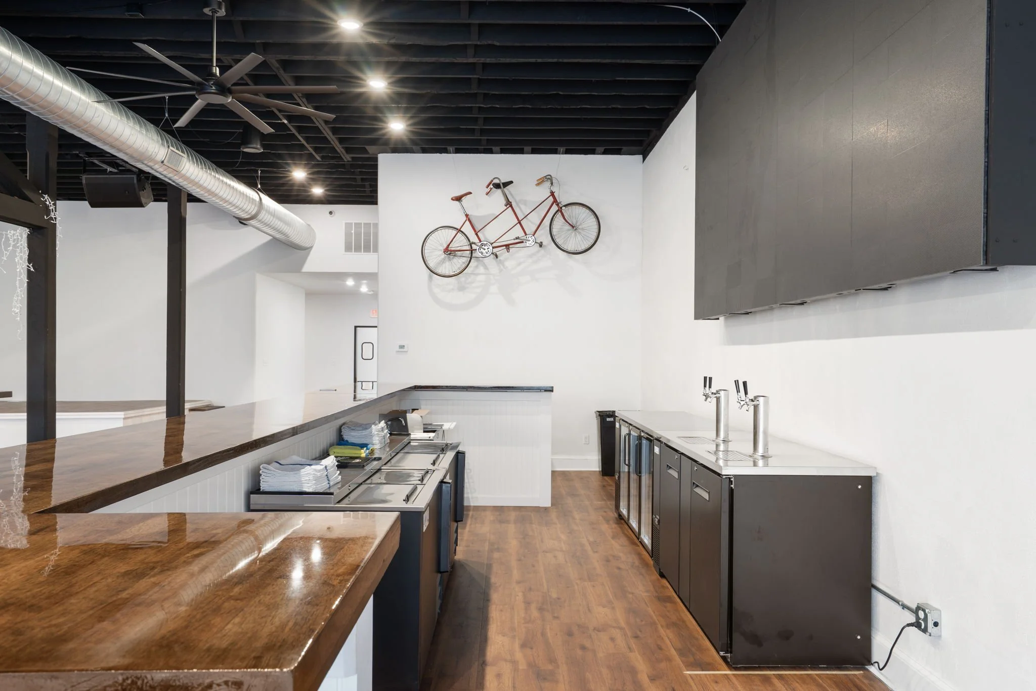 Interior of a modern bar or kitchen area with a wooden counter, black cabinets, and a white wall decorated with a vintage bicycle. Dishes and towels are on the counter, with ceiling fans and industrial ductwork overhead.