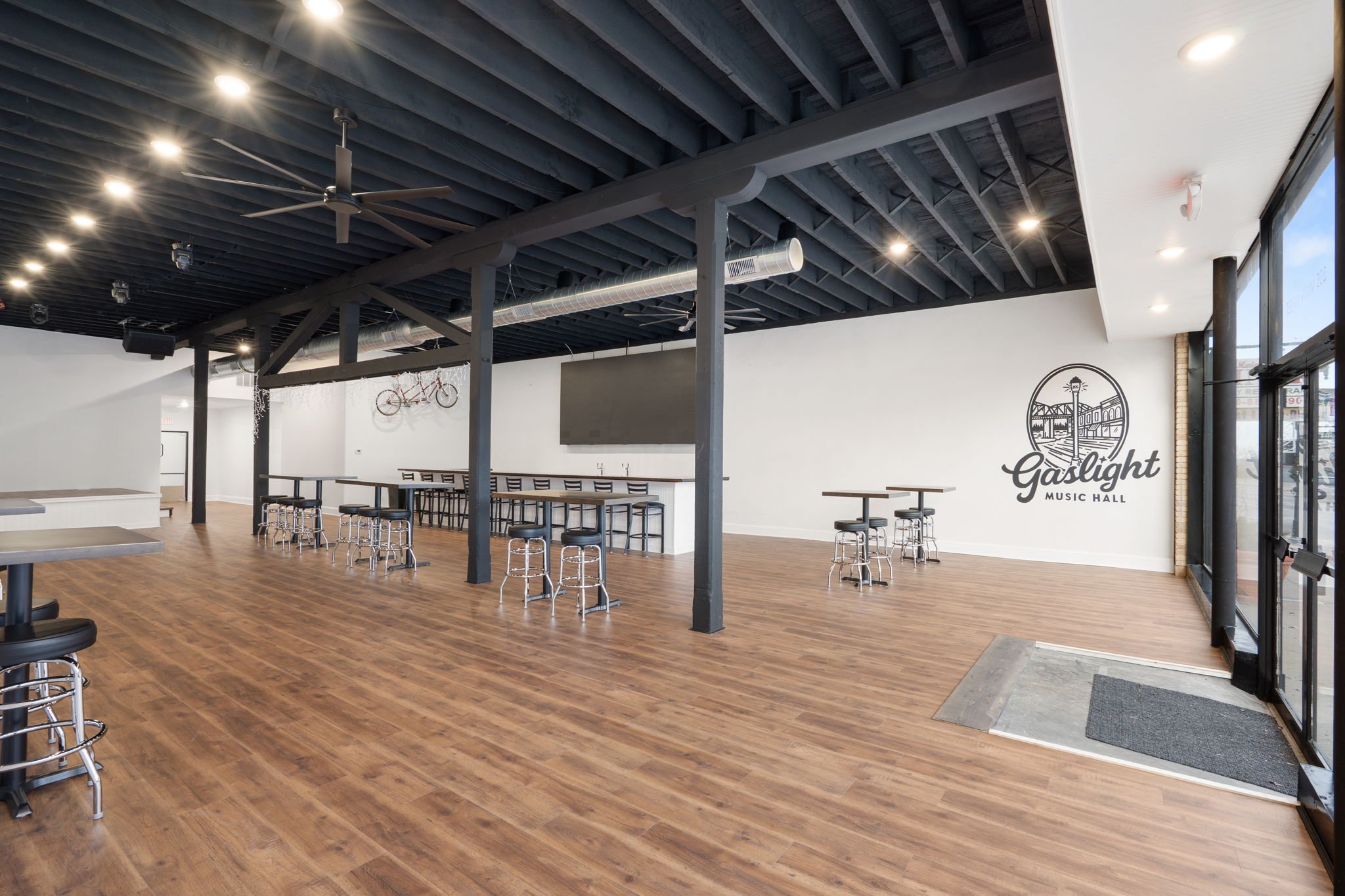 Empty interior of Gaslight Music Hall with wooden floors, black ceiling, and wall logo, featuring tables, chairs, bar-height stools, and large windows.