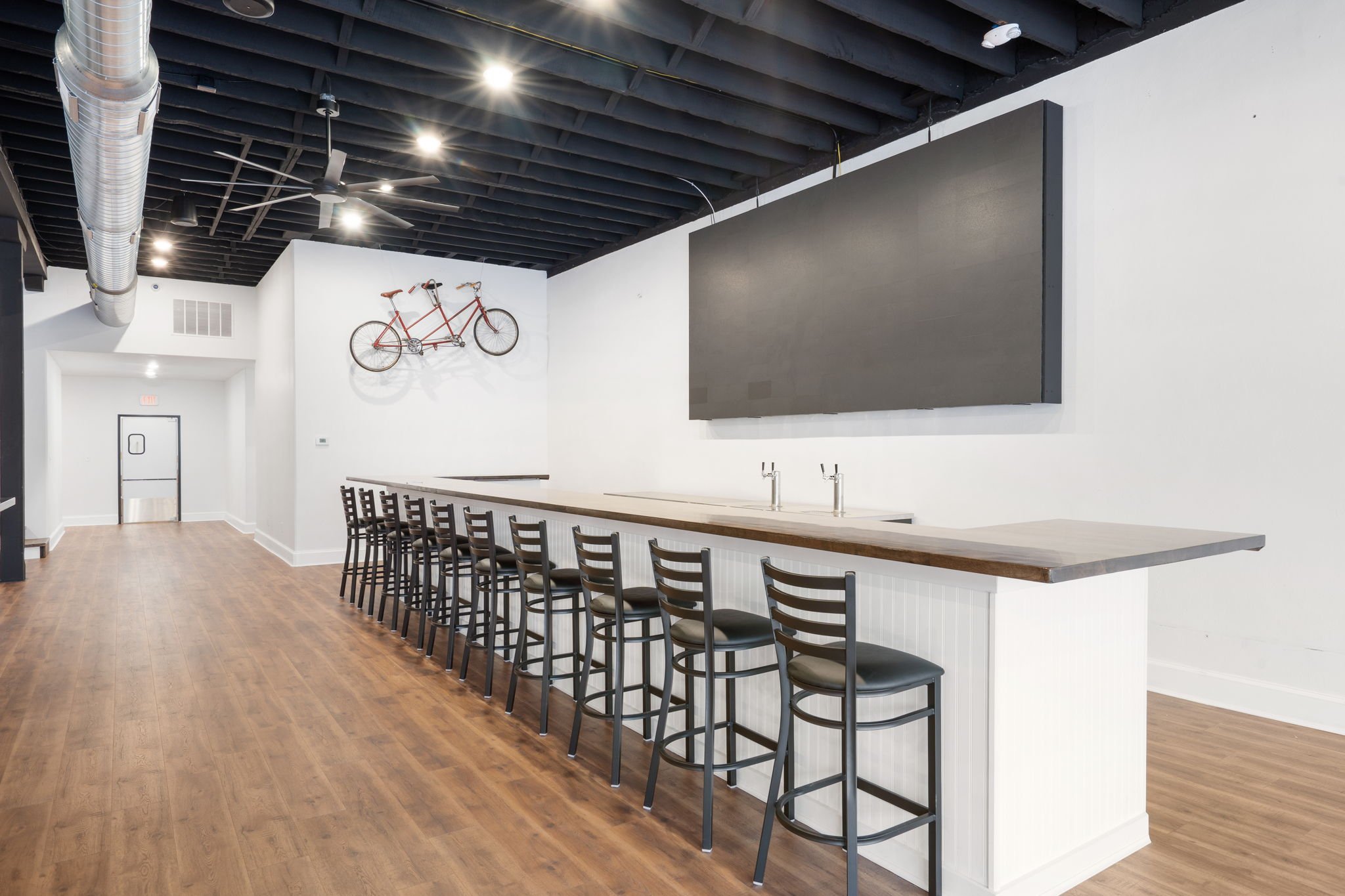 Interior view of a modern bar or cafe with a long counter and multiple black chairs, white walls, and a wooden floor. There is a bicycle mounted on the wall and a large black screen or panel above the counter. The ceiling has exposed black beams and 