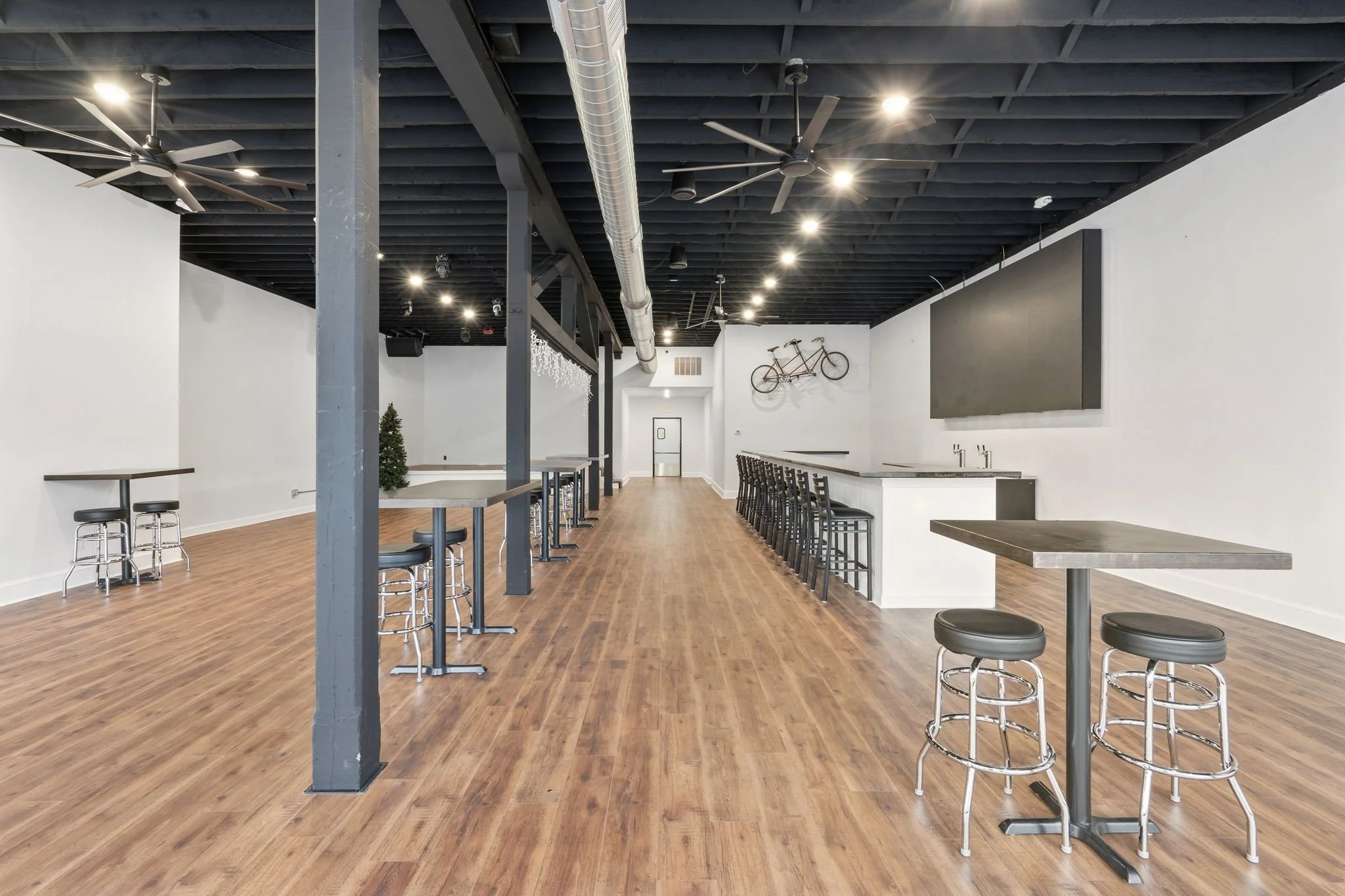 Empty bar with high stools, and high tables on a wooden floor, industrial style with exposed ductwork and ceiling fans, wall art including a bicycle, and a large black rectangular screen.