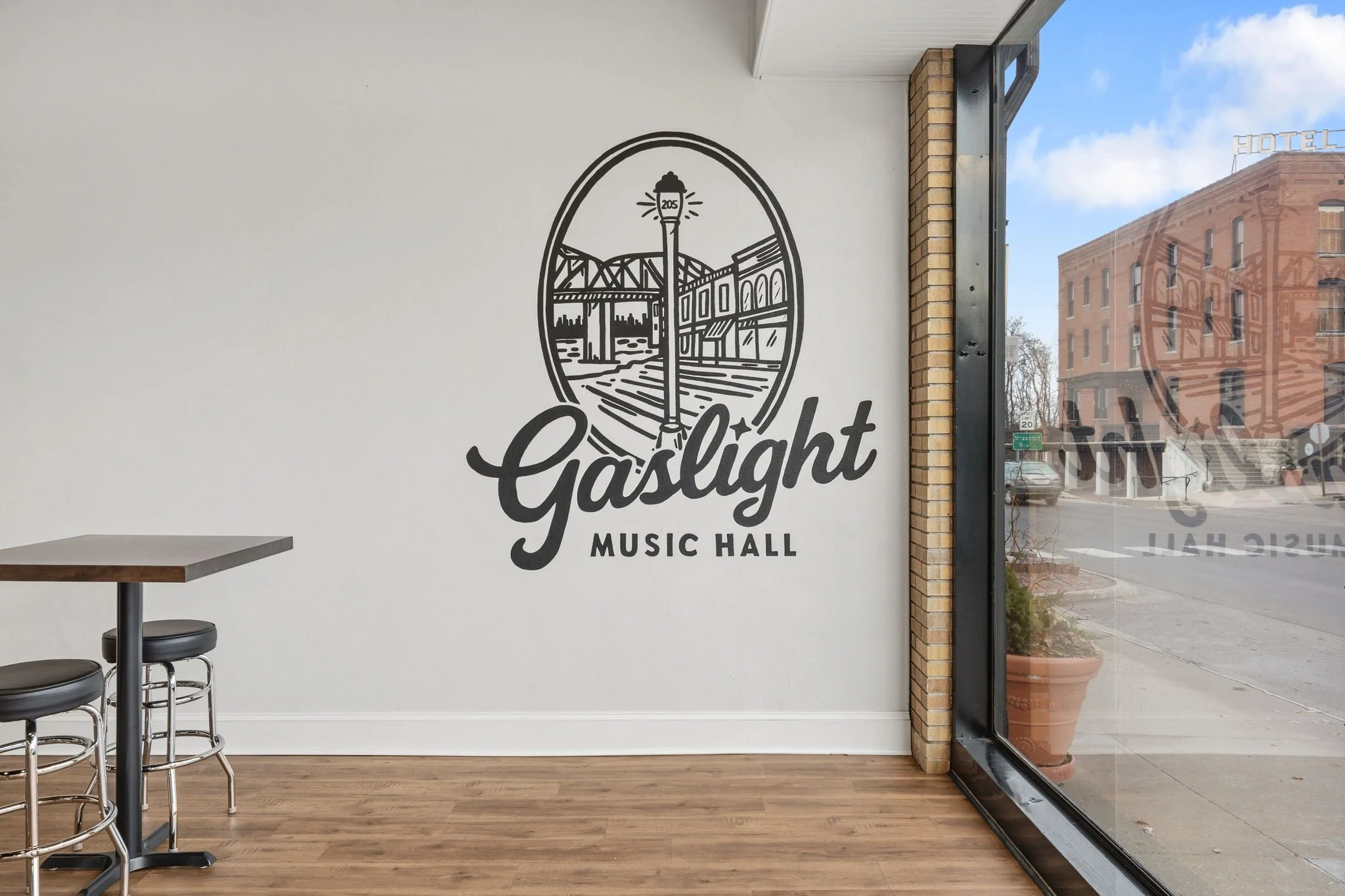 Interior of Gaslight Music Hall with a white wall, wall art of a mountain with a lighthouse and bridge, and a large window showing a street scene outside.